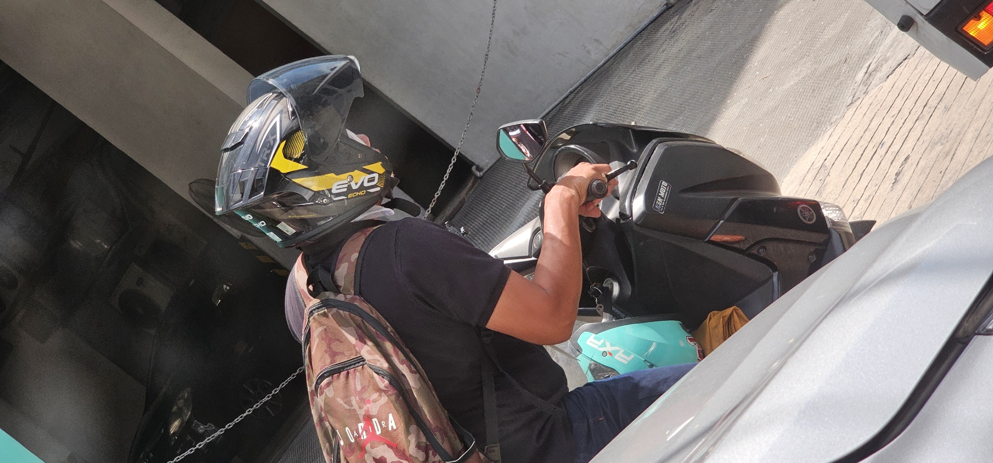 A person wearing a black shirt, blue jeans, and a black and yellow helmet is riding a scooter. The person is carrying a camouflage backpack. The scooter is black with a teal helmet and a yellow object on the back. The scene appears to be on a street with a gray concrete ground. The background shows a dark structure with metal chains. The image is taken outdoors, during the day, with bright sunlight.