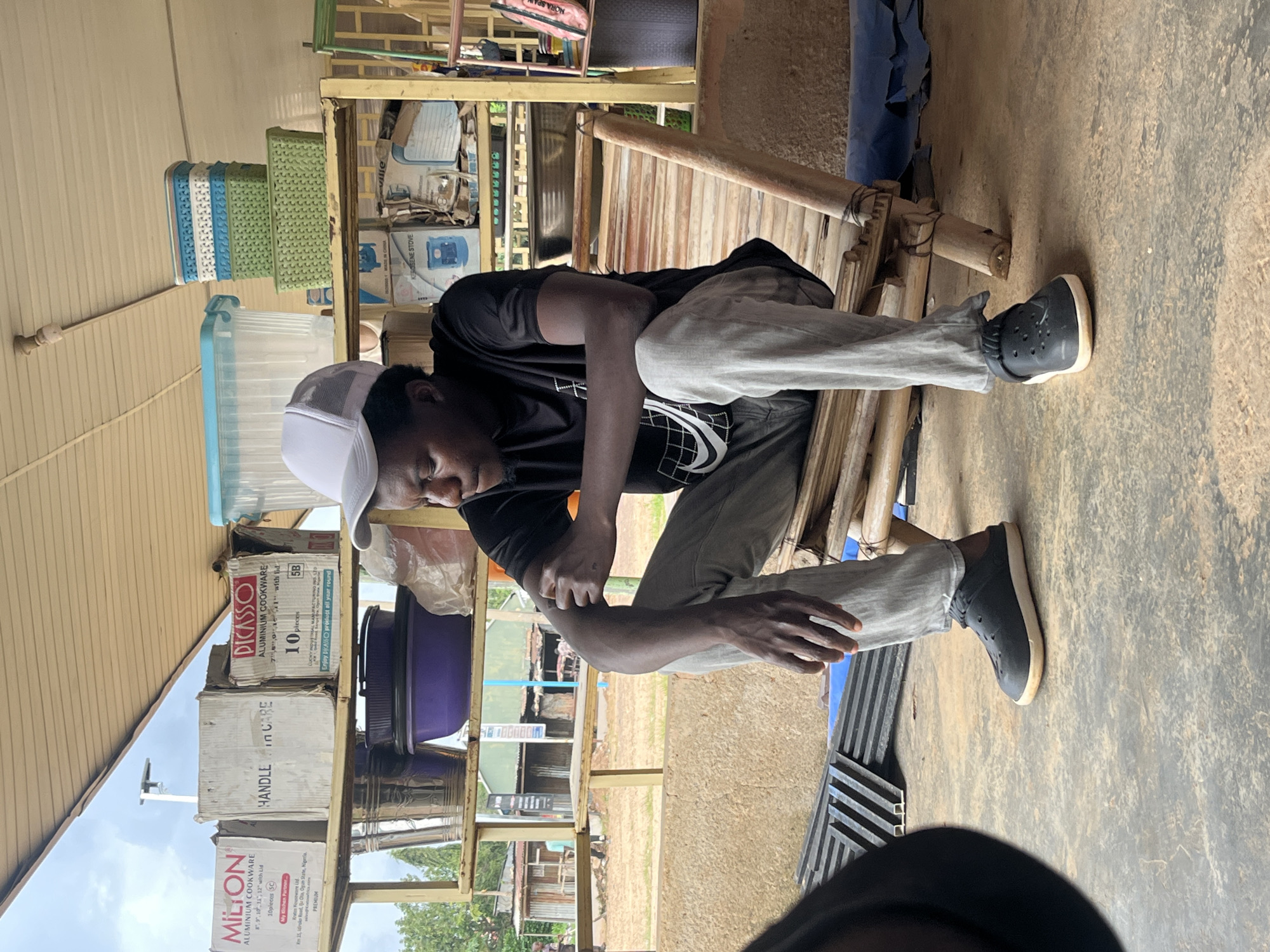 A young Black male, wearing a white baseball cap, black t-shirt with a white graphic design, and light grey pants, sits on a bamboo chair in what appears to be an outdoor market or shop. He is positioned centrally in the frame, facing to his left, with his arms resting on his legs and his hands clasped. The background features shelves stocked with various items, including boxes of cookware, plastic containers, and baskets. Beyond the shelves, a glimpse of an outdoor scene with buildings and trees is visible, suggesting an urban or semi-urban environment. The ground is concrete, and there are some black metal grates on the left side of the image. The overall lighting indicates it is daytime, possibly overcast.