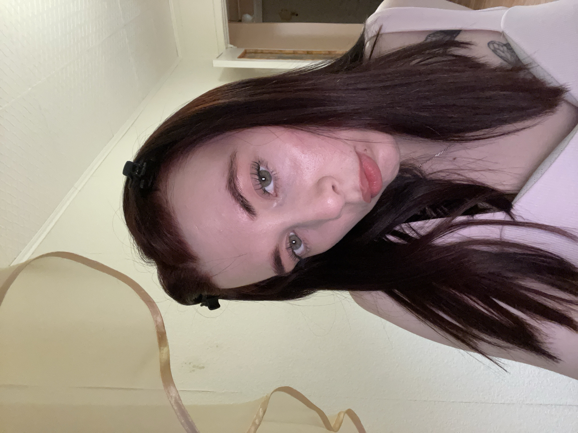 A young woman with dark auburn hair and pale skin is positioned in the frame, looking directly at the camera. A small black hair clip secures the hair at the left side of her face. She has light eyes and a pursed expression. A silver necklace is visible around her neck. A tattoo is visible on the upper right shoulder. The background features a cream-colored wall, a sheer curtain in the lower left corner, and a window above and behind the woman, partially obscured by her hair. The image provides no information to suggest a location in the city of Chaikovsky, Russia.