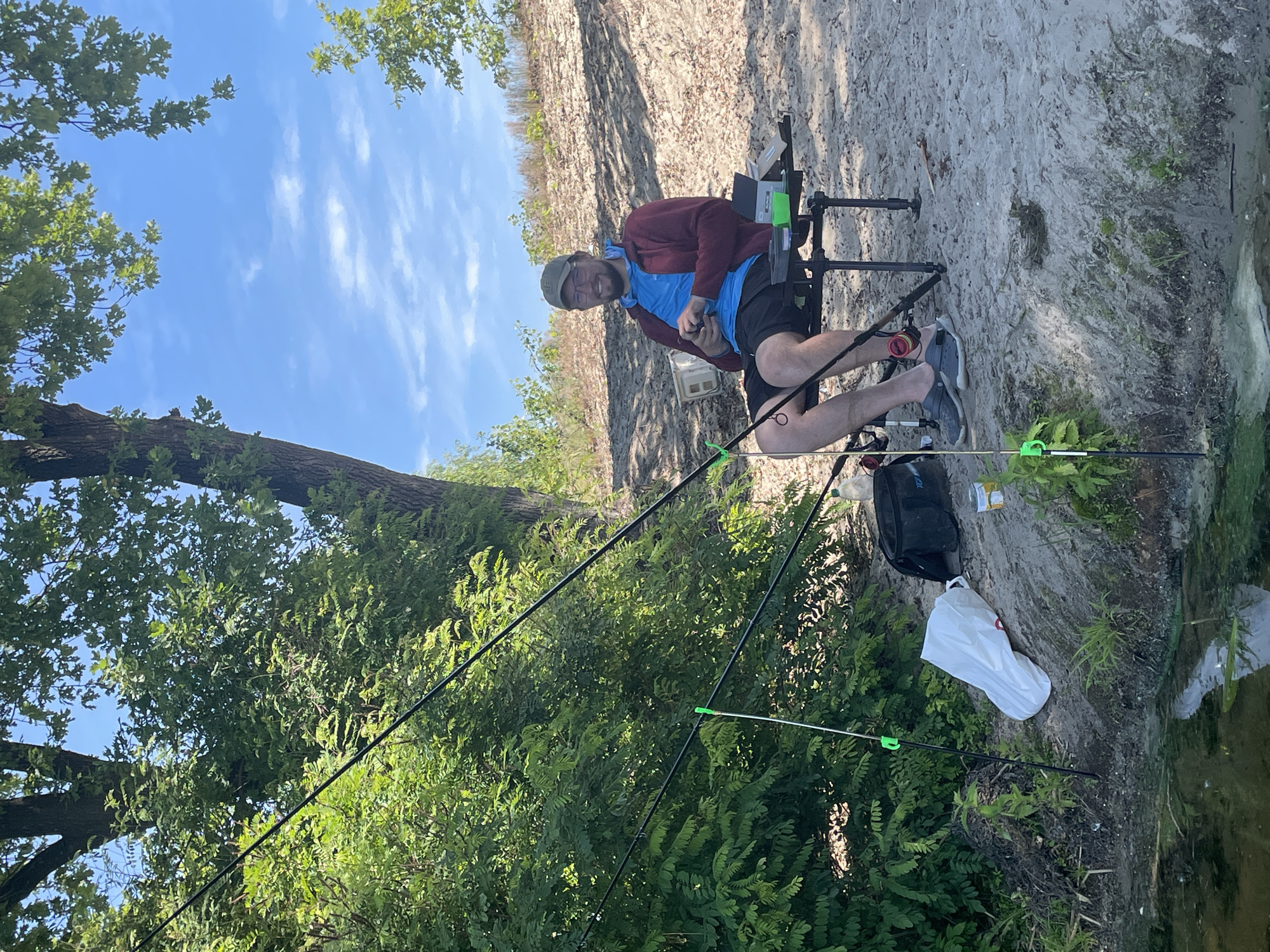 A man is fishing on a sunny day near the Dnipro River in Kyiv, Ukraine. He is sitting in a portable fishing chair on a sandy bank, with fishing rods set up in front of him. He is wearing a blue t-shirt, a maroon jacket, black shorts, a baseball cap, and glasses. He is smiling and holding a fishing reel. Several pieces of fishing equipment, including a tackle box and a bait container, are visible around his chair. A white plastic bag and a black fishing bag are also on the bank near him. The background consists of lush green trees and a clear blue sky with a few scattered clouds. The Dnipro River is visible in the foreground, with some aquatic plants on its surface.