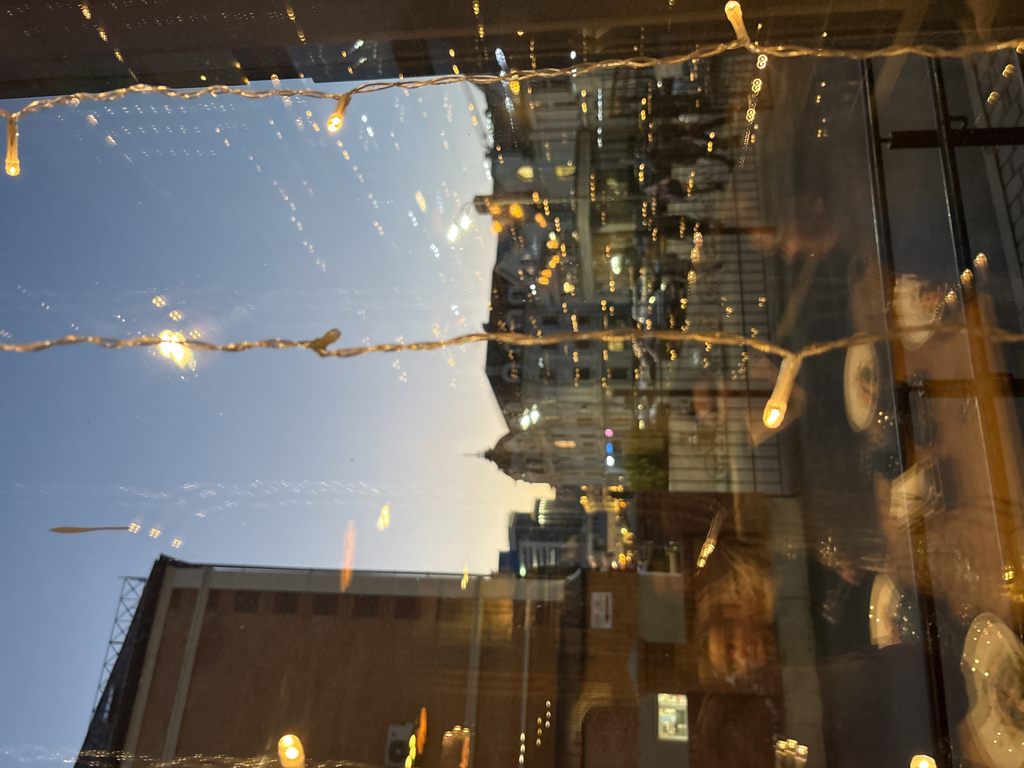 The image shows a view of Kazan, Russia, at dusk, seen through a window decorated with fairy lights. The window reflects the interior of a building, possibly a restaurant, showing blurred figures of people sitting at tables with plates. Outside, a brick building is visible below the skyline, which includes a domed structure and various buildings illuminated with lights. The sky is a gradient of blue to orange.