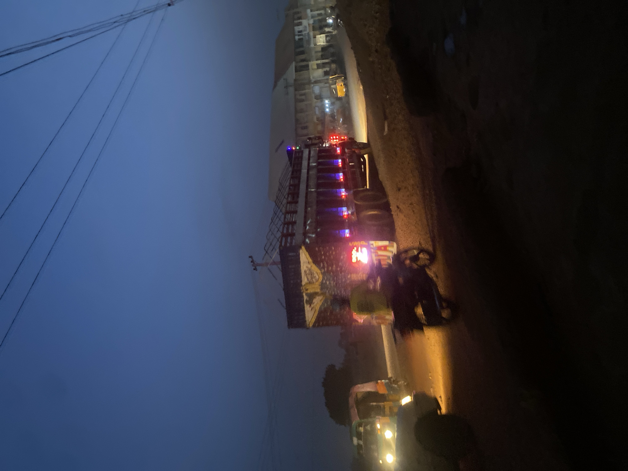 The image depicts a street scene at dusk or night in Tudun Wada, Nigeria. A large truck with multi-colored lights and possibly an overloaded cargo is parked along the side of the road. A motorcycle and an auto rickshaw are also visible on the street, their headlights illuminating the immediate area. A person is visible near the motorcycle, slightly blurred. Buildings line the opposite side of the road from the truck. Overhead power lines are visible against the dim sky.