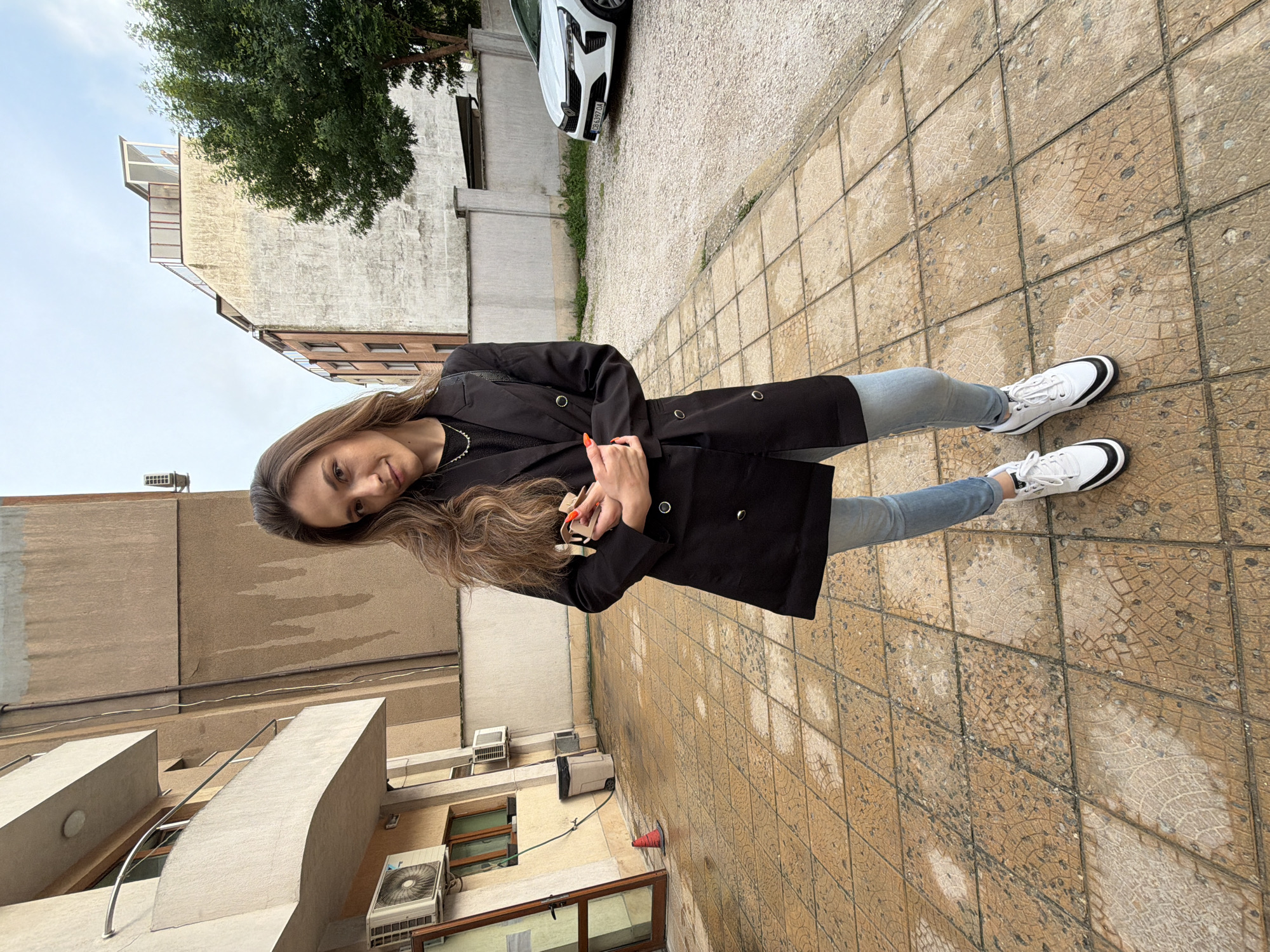 A young woman stands outdoors in an urban environment, posing for the camera with a gentle smile. She has long, wavy brown hair and is wearing a black double-breasted coat over a dark top, light blue jeans, and white sneakers with black accents. Her fingernails are painted orange, and she holds a small object, possibly keys or a phone, in her hands.
The setting appears to be a paved area next to buildings. The ground beneath her is made of light brown/beige tiles with a textured, slightly weathered pattern. Behind her, to the left, is a multi-story building with air conditioning units and windows. Further back and to her right, another building with balconies and a lighter-colored upper section is visible, with a large green tree growing nearby.
To her right, a white car is parked on a patch of gravel, with its front fascia and the Renault logo clearly visible. The license plate "B 4397 OA" is legible. In the background on the right, near a building, a white generator and a small orange traffic cone are present. The lighting suggests it is daytime, possibly overcast, providing soft, even illumination.