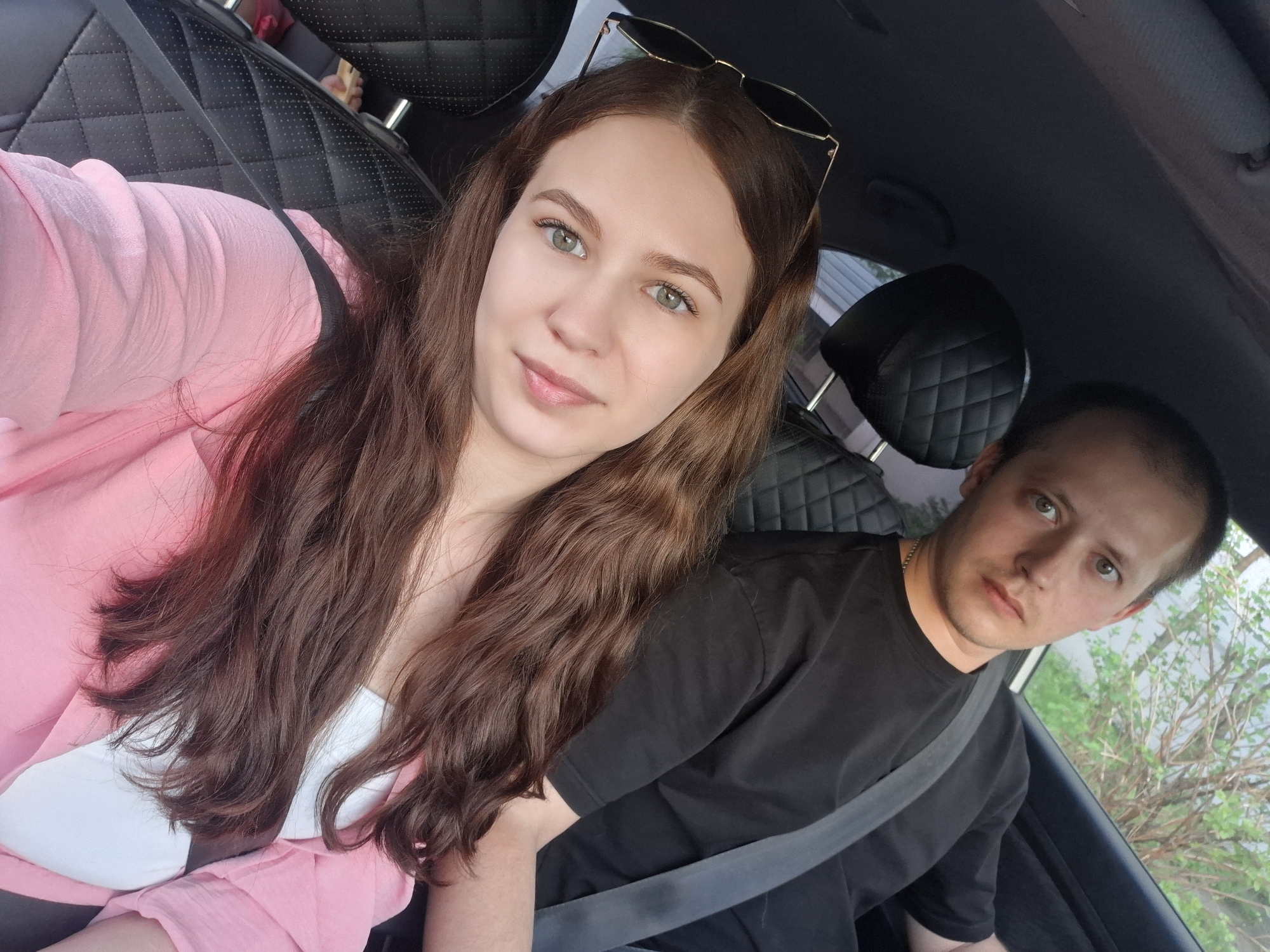 A medium shot captures a woman and a man inside a car. The woman, positioned to the left, has long, brown hair and is wearing a pink top. Sunglasses are perched on her head. The man, situated to the right, wears a black t-shirt and a seatbelt. The car's interior is visible, including the leather seats. Outside the car window, a green bush and glimpses of the surroundings are seen. The location is Chaikovsky, Russia.