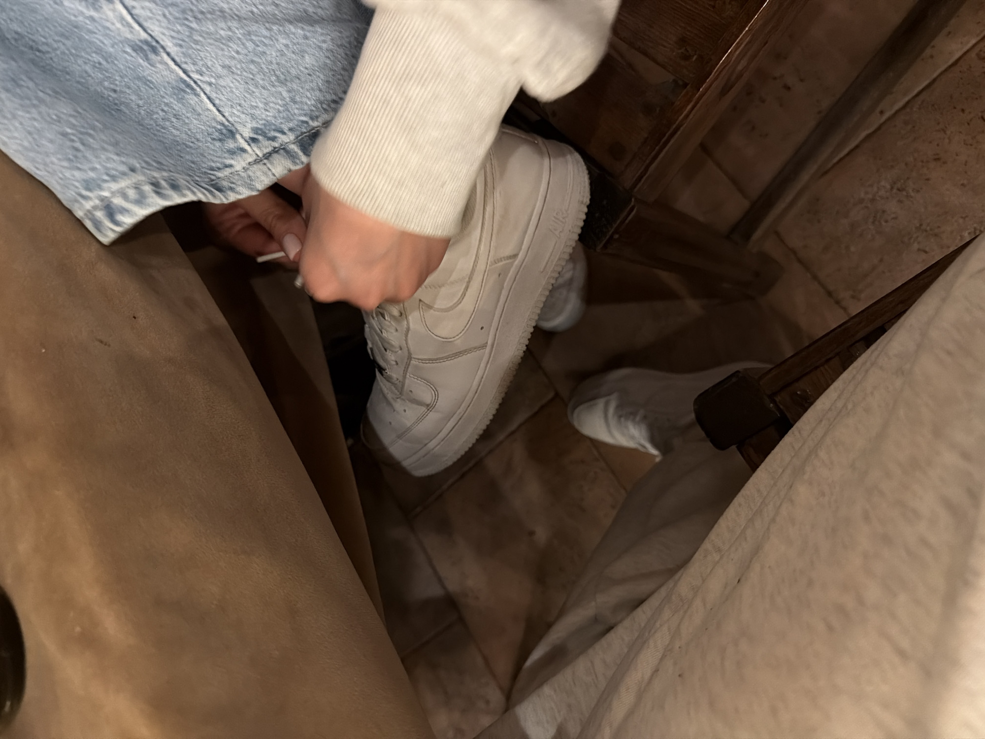 The image presents a close-up, downward view of an indoor setting, likely in Yerevan, Armenia. The central focus is a person's hand, adorned with light pink nail polish, in the process of tying the white laces of a white Nike Air Force 1 sneaker. The individual is wearing light blue denim jeans and an off-white, ribbed long-sleeved top, whose cuff is visible. The word "AIR" is discernibly embossed on the sole of the sneaker.
In the foreground, a warm brown fabric, possibly a pant leg or seating, is visible on the left. To the right, another person's leg, dressed in light beige or cream-colored fabric, is partially seen, along with a second white sneaker further back. The floor is covered with light brown or beige tiles, showing distinct grout lines and some minor wear or dirt. Dark wooden furniture, appearing to be chair or table legs, is visible in the upper and mid-right sections of the frame. The lighting is dim and artificial, indicating an indoor environment, possibly in the evening or at night. The scene depicts a casual moment, perhaps in a home or a relaxed social space.
