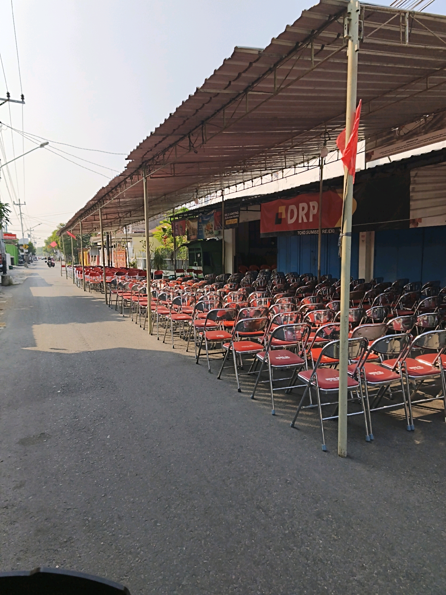 The image shows a street in Surakarta, Indonesia, lined with rows of red chairs under a temporary canopy. The street appears to be in front of shops, with blue doors and a banner with the word "DRP" visible. The canopy is supported by poles, and a red flag is attached to one of them. There are no people visible in the image, but the presence of chairs suggests a gathering or event is planned or has recently occurred. The sky is clear and the lighting indicates it is daytime.