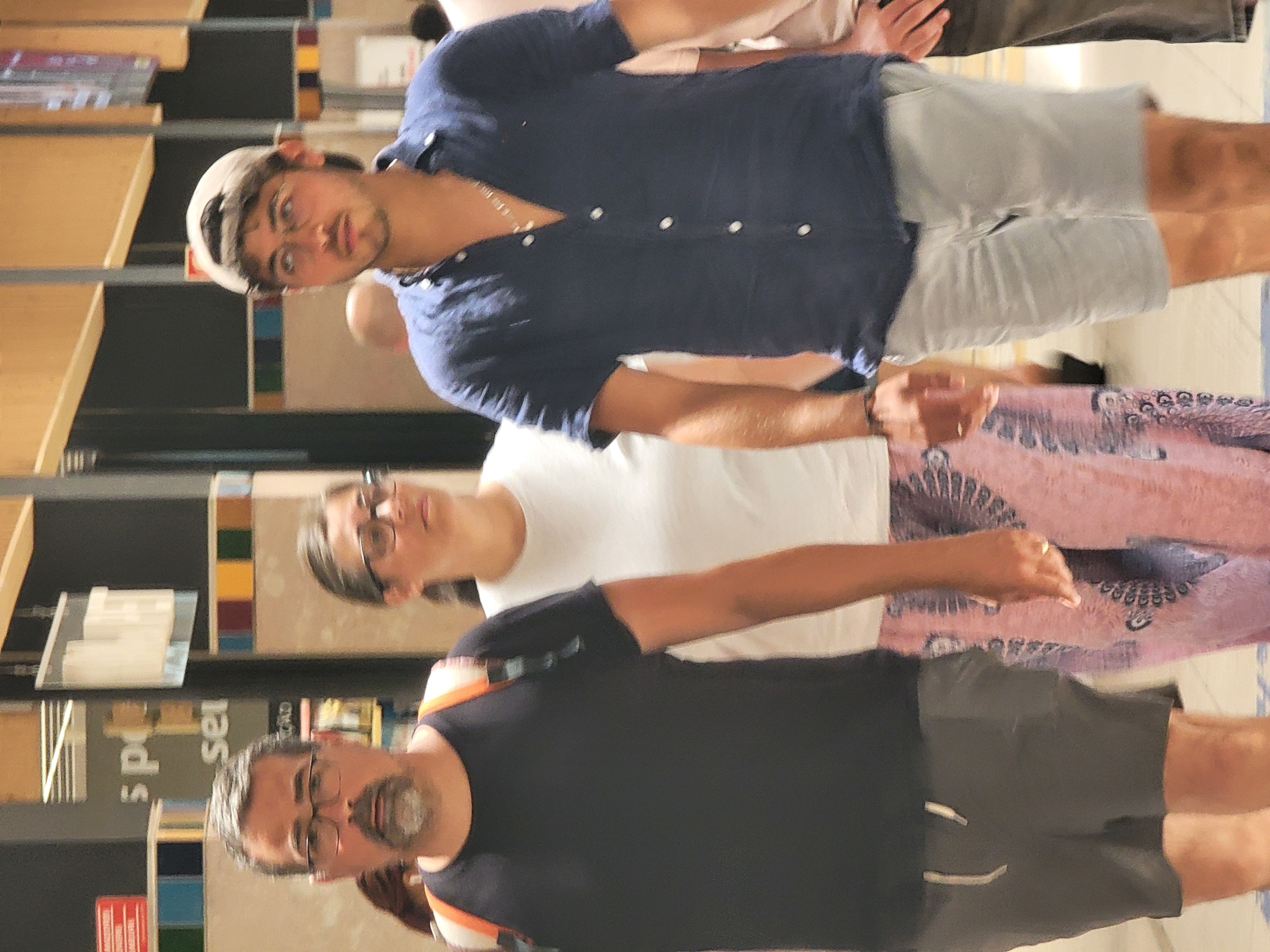 Three individuals are walking side-by-side in what appears to be an indoor public space, possibly a shopping center or airport terminal, with vertical beige architectural elements and signage visible in the background. The person on the left is a man with glasses, a goatee, and graying hair, wearing a black t-shirt and grey shorts, with an orange backpack slung over his shoulders. To his right is a woman wearing glasses and a white top, with her arm around the man's shoulder, and a patterned pink skirt. On the far right is a young man wearing a blue button-down shirt, grey shorts, and a baseball cap. The lighting suggests an indoor environment, and the overall scene depicts casual movement. The setting, with specific architectural details and visible signage that hints at commercial or transportation hubs, is consistent with locations in Faro, Portugal.