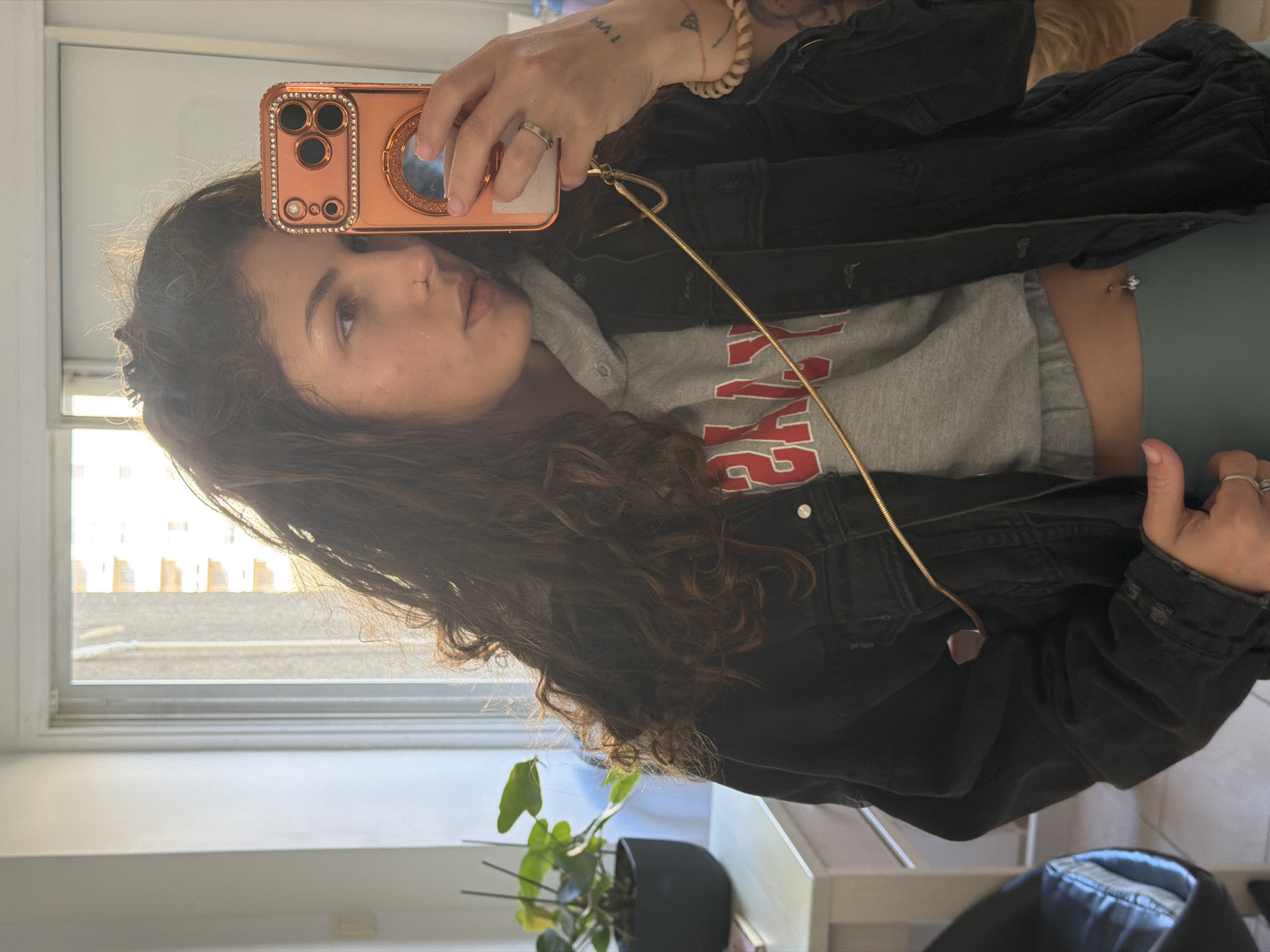 A young woman with curly brown hair takes a mirror selfie indoors. She is wearing a grey sweatshirt with red lettering, a black denim jacket, and green pants. She is holding a rose gold-colored phone with a jeweled case. Tattoos are visible on her hand and arm. A window with white trim is in the background. A potted plant sits on a table near the window. A glimpse of furniture, including a chair, is also visible. The image was taken inside a room located in Kiryat Bialik, Israel.
