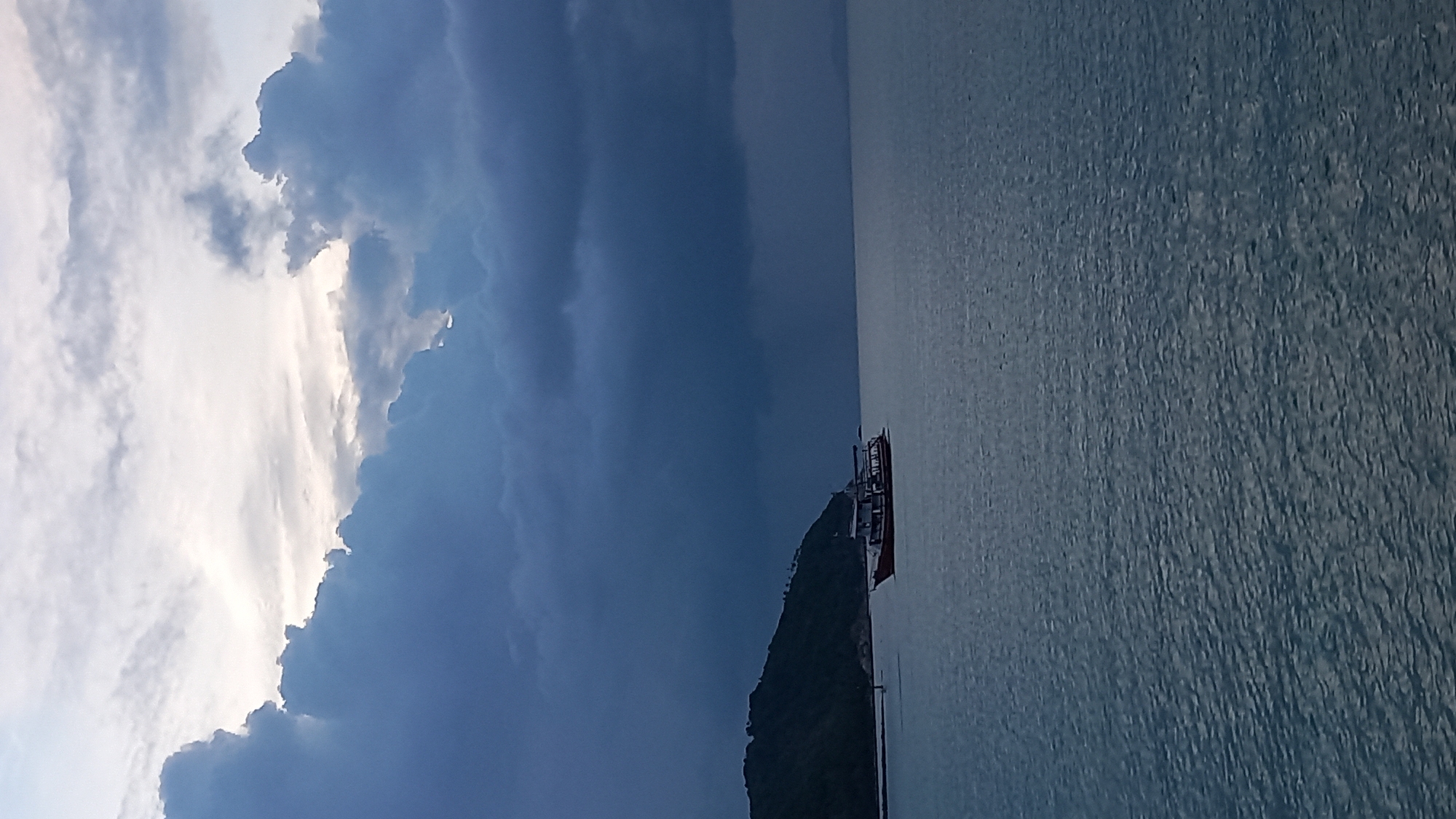 A boat sails on the sea in Ko Pha-ngan District, Thailand, under a cloudy sky. The boat is red and white and has a dark roof. A green, hilly island is visible on the left side of the image, with some trees on its slopes. The water is choppy and reflects the gray sky. The clouds are dark and heavy, with some patches of lighter sky showing through. The overall mood of the image is dramatic and somewhat ominous.