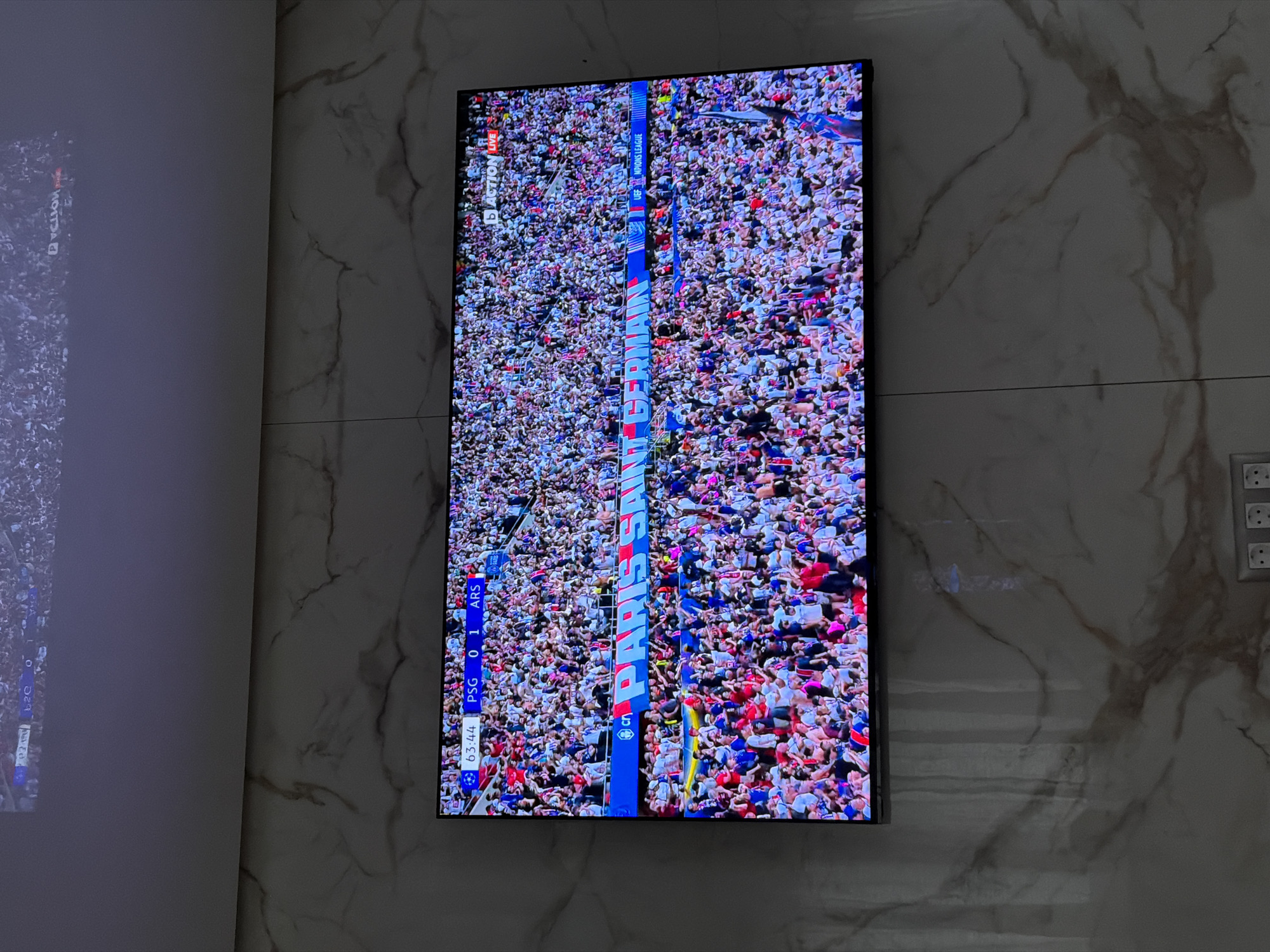 The image shows a TV screen displaying a football (soccer) match between PSG and Arsenal. The score is PSG 0, Arsenal 1, at the 63:44 minute mark. The screen is mounted on a light-colored, marble-patterned wall. A blurred screen can be seen on the left side of the frame. Electrical sockets are visible on the right side of the frame. The image was captured indoors. The location in the city of Asenovgrad, Bulgaria, cannot be determined from the image.
