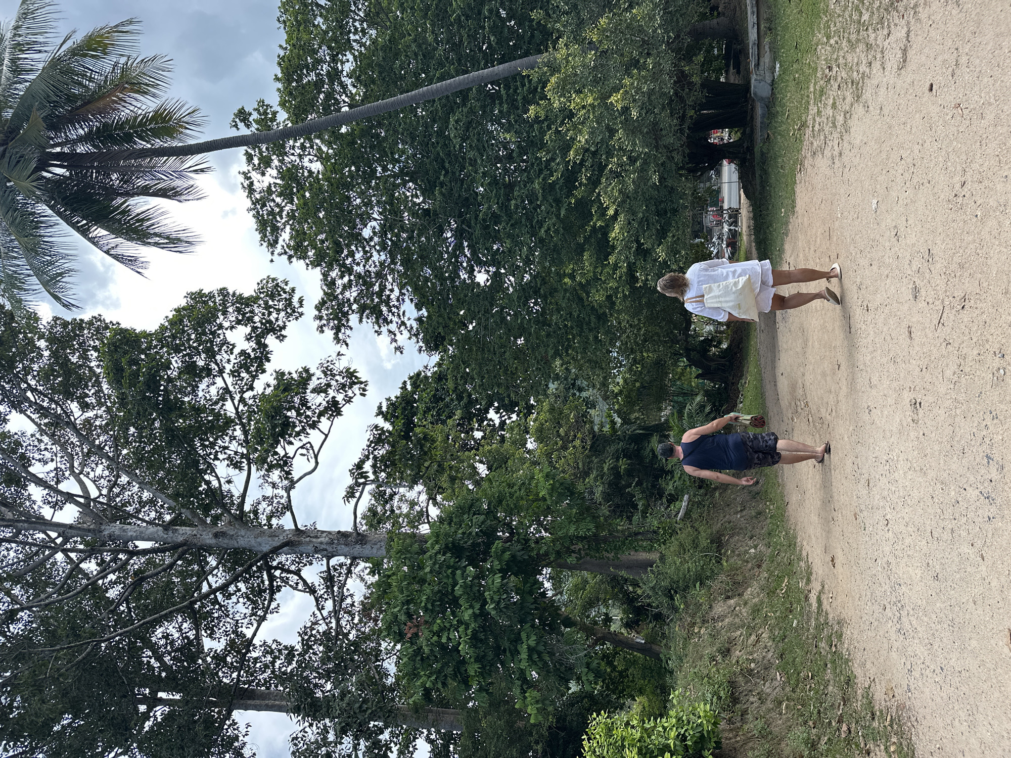A man and a woman are walking away from the camera on a dirt path in a tropical environment, likely on the island of Ko Samui District, Thailand. The man, on the left, wears a dark blue tank top and patterned shorts, carrying a light-colored bag over his right shoulder. The woman, on the right, is dressed in a white short-sleeved shirt and white shorts, and carries a large white tote bag over her left shoulder.
The path is wide and unpaved, with loose gravel and dirt. Lush green vegetation lines the sides of the path, with large trees and palm trees dominating the background. The sky is overcast with some visible clouds. In the distance, through the trees on the right, hints of buildings and parked motorbikes suggest a more developed area beyond the path. The overall scene depicts a casual stroll through a verdant, possibly rural or semi-rural, part of the island.