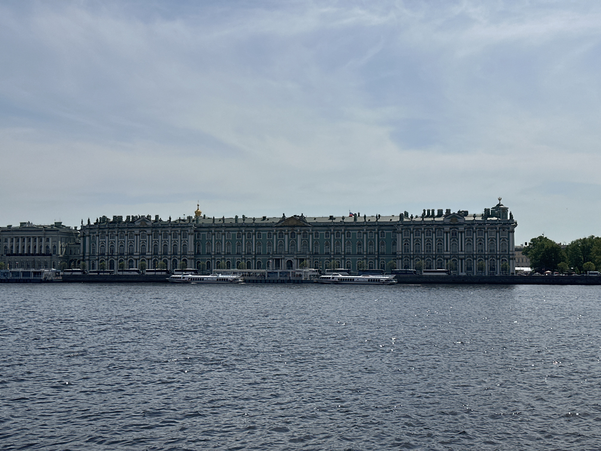 The image depicts the Winter Palace, a prominent landmark located in Saint Petersburg, Russia, situated on the bank of the Neva River. The grand facade of the palace extends horizontally across the frame. Several white boats are docked along the riverbank in front of the palace. The Neva River fills the foreground, exhibiting rippled water with sunlight reflecting off its surface. The sky above is overcast with scattered clouds. The overall scene presents a tranquil view of this historic location.
