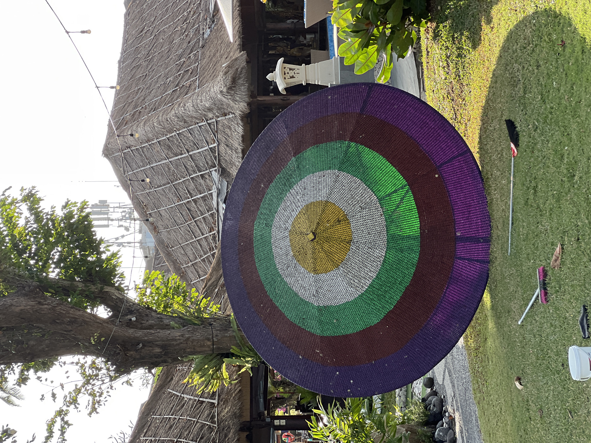In Denpasar, Indonesia, a large, circular, woven target with concentric rings of purple, maroon, green, white, and yellow stands upright on a grassy lawn. Behind the target, traditional thatched-roof buildings and a mature tree with visible aerial roots are visible. The scene appears to be outdoors, with sunlight casting shadows on the grass. A broom and a white bucket are present on the lawn in the foreground. The background shows more foliage, a stone pathway, and a white decorative lantern near a swimming pool. Cell towers are visible in the far distance, suggesting an urban or semi-urban environment.