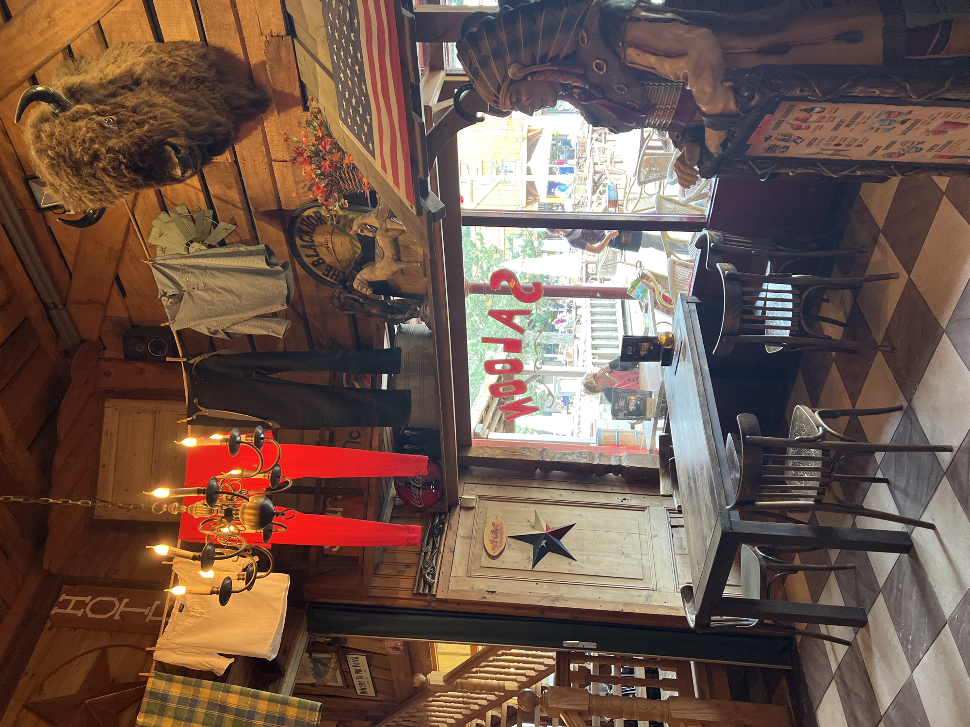The image shows the interior of a rustic-themed establishment, possibly a restaurant or bar, located in Collendoorn, Netherlands. The walls and ceiling are constructed from wood, creating a warm and country-style atmosphere. In the foreground, a large wooden table with chairs is set on a checkered floor. To the right, a life-sized statue of a Native American chief stands prominently. Above the table, a chandelier with multiple candle-shaped lights hangs from the ceiling. Laundry, including a white shirt, red pants, and dark trousers, is hung on a clothesline across the upper part of the room. Mounted on the wall is a taxidermied bison head, adding to the wild west theme. Behind the table, a large window looks out onto an outdoor seating area with tables and umbrellas, and further beyond, buildings typical of a village or tourist attraction. On the window, the word "SALOON" is written in large, red lettering. The overall impression is of a themed establishment aiming for a nostalgic or adventurous ambiance.