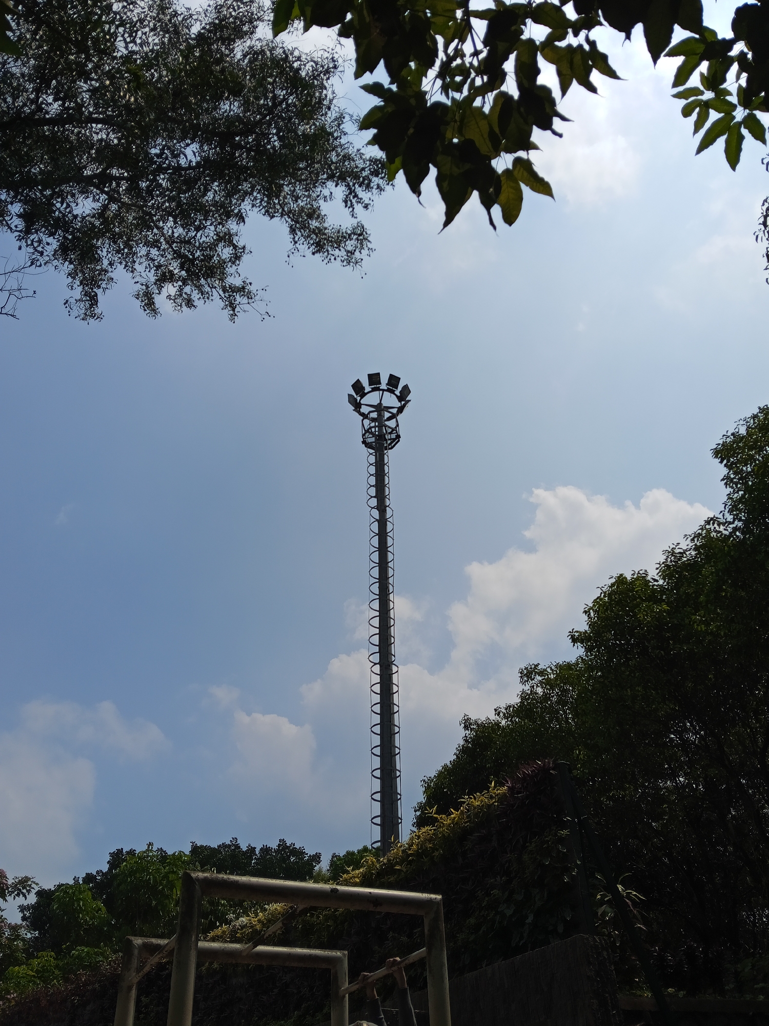 A tall, metallic, floodlight tower stands against a bright blue sky, punctuated by scattered white clouds. The tower, with a spiral staircase visible along its length, is topped by a circular array of multiple spotlights. Silhouetted against the sky, trees with dense foliage frame the scene from the top and sides. In the foreground, part of a metal structure, possibly part of a sports facility, is visible. The environment appears to be an outdoor space, possibly a park or a sports field in the city of Semarang, Indonesia, given the vegetation and the lighting tower which is common in such locations.