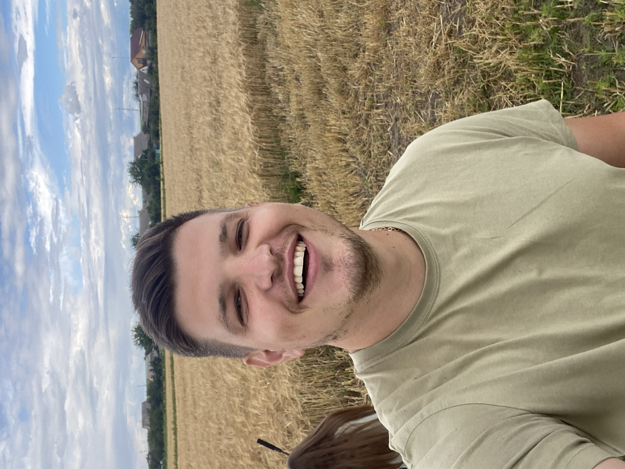A man is taking a selfie in a wheat field, with a person's back to the camera in the background. The man is smiling and wearing a light green t-shirt and a gold chain. Behind the field, a village with houses and trees is visible under a cloudy sky. The location is Yablunivka, Ukraine.