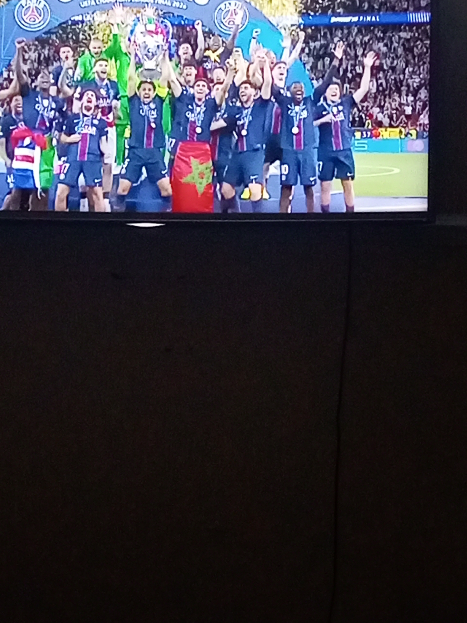 The image shows a television displaying a group of people, presumably a soccer team, celebrating a victory. They are holding a trophy and flags, one of which appears to be the flag of Morocco. The backdrop features a crowd of spectators, suggesting a large stadium or event. The text "UEFA CHAMPIONS LEAGUE FINAL 2020" is visible, indicating the event's name and year. The TV is mounted on a wall. The location is Unknown, Unknown.