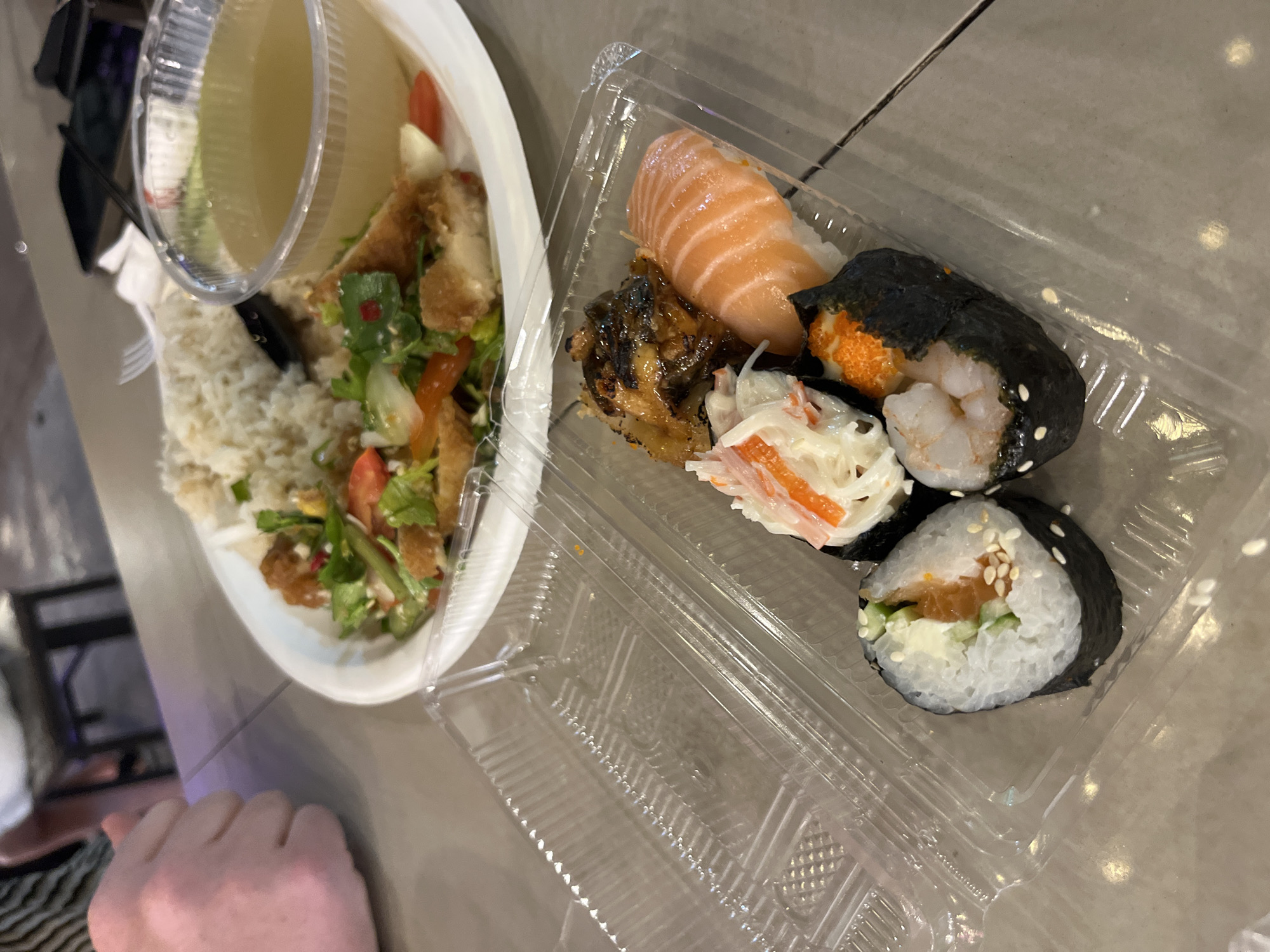 The image shows a take-away meal consisting of a salad with rice and a small container of dressing, alongside a plastic container of sushi. The sushi selection includes nigiri (salmon and eel), a type of sushi roll with shrimp and roe wrapped in nori, and a California roll (imitation crab). The scene appears to be indoors, possibly a home or hotel room, based on the smooth, neutral-toned surface the food containers rest on. The presence of an arm/hand in the lower left corner indicates a person is nearby. The photo was taken in Pattaya City, Thailand.