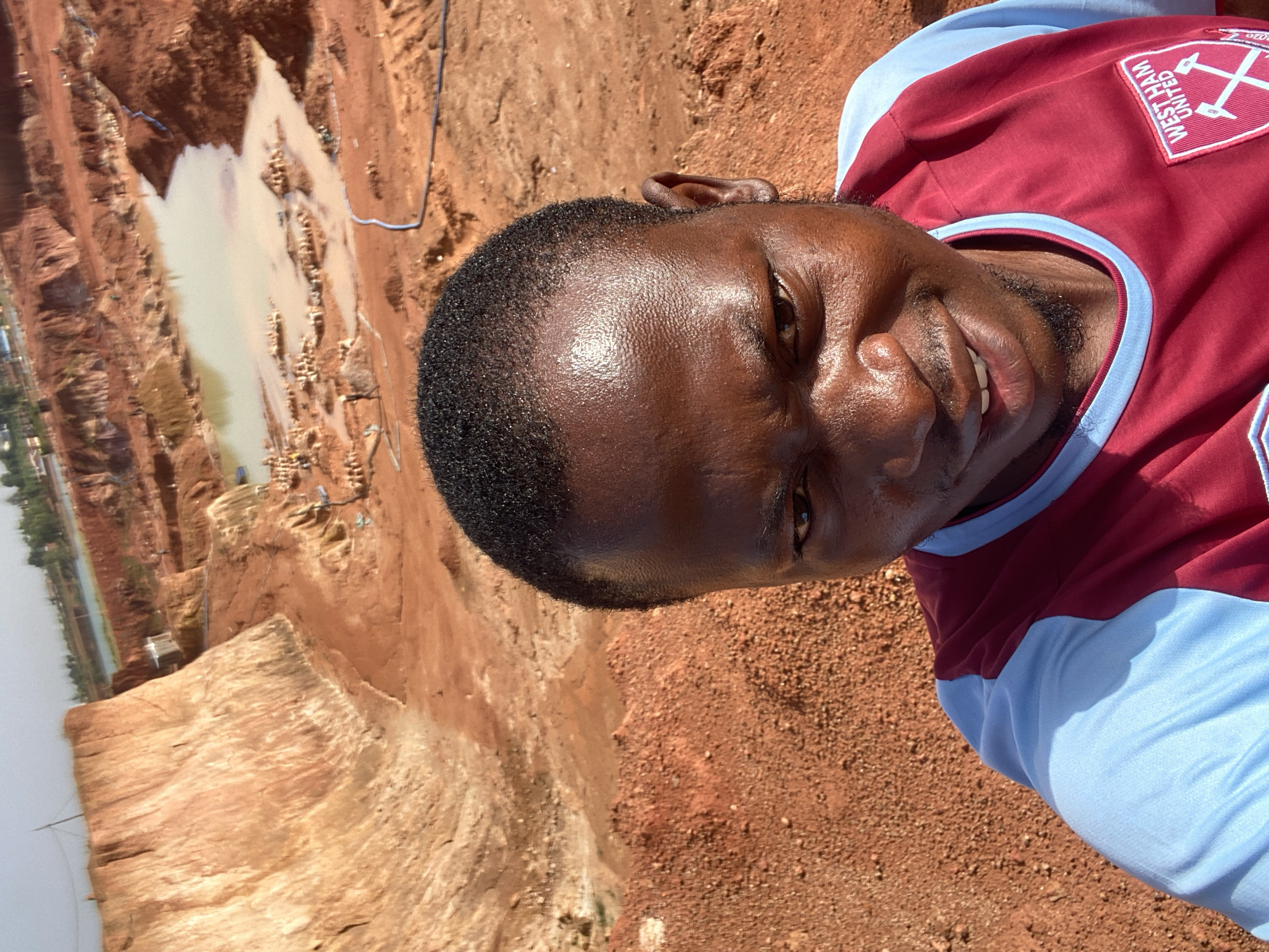 A close-up captures a man in a West Ham United jersey against a backdrop of a construction site. The man, facing the camera, is positioned in front of a deep excavation. The construction site features reddish-brown soil, and a pool of muddy water is visible in the distance. The location is the city of Unknown, Unknown.