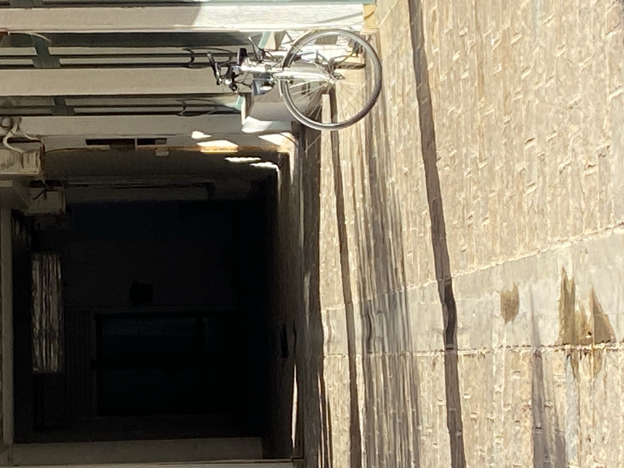 The image shows a bicycle hanging upside down from a ceiling or structure above a dark passage or shaft. The bicycle is silver and has a road bike-style frame. The ceiling above appears to be made of concrete or a similar material, with metal supports. Below, the shaft is dark and deep, possibly a service access point, an elevator shaft, or a stairwell entrance. The adjacent wall is constructed of stone or concrete blocks with a textured surface.
The photo was taken looking up from below, causing the skewed perspective. There are no people visible in the image. The location of the photo is in Budva, Montenegro.
