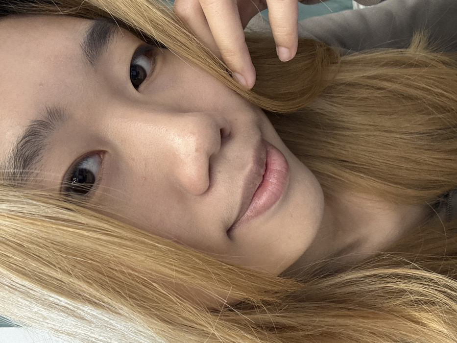 The image presents a close-up, frontal portrait of an individual of East Asian descent, likely female, situated indoors. The subject has light blonde or light brown straight hair that frames her face, dark eyes directly engaging the viewer, and dark, defined eyebrows. A subtle, closed-mouth smile is evident on her lips. Her left hand, with visible fingers, rests lightly against her hair on the right side of the frame. A portion of a grey garment is visible at the bottom right. The background is blurred, showing a light-colored surface to the upper left and a darker, possibly teal or blue-green, surface to the upper right and far right. The specific location within the city of Nan'an District, China, cannot be determined from the visual information provided.