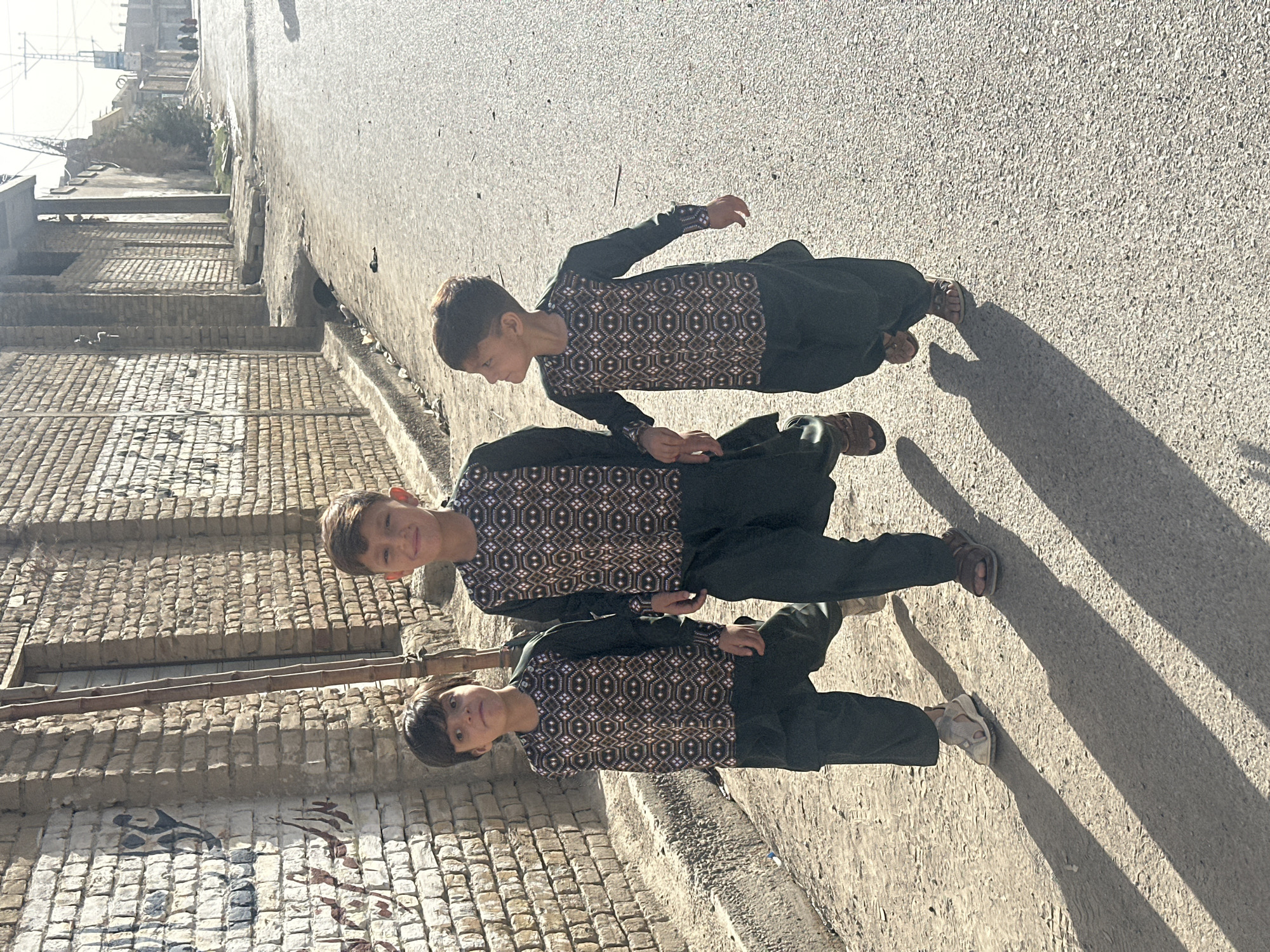 Three boys in matching brown patterned long-sleeved shirts and dark pants are standing on a street in Quetta City Tehsil, Pakistan. They are lined up behind each other, with the youngest boy in front and the oldest in the back. The boys are holding hands. A brick building is to the left. The street is asphalt. Shadows of the boys are cast to the right.