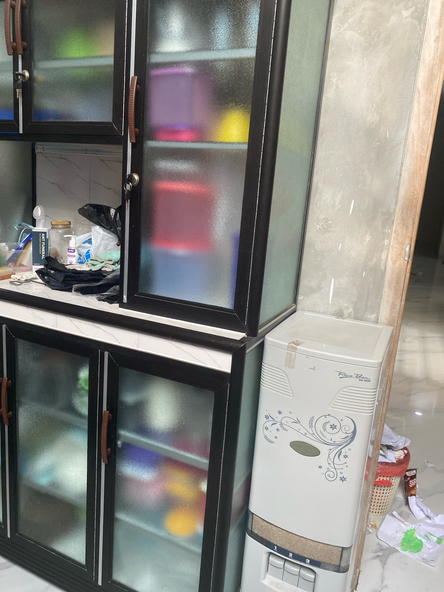 The image shows a corner of a room, possibly a kitchen or storage area, with a large cabinet and a rice dispenser. The cabinet is dark-framed with frosted glass doors, revealing various items inside, including colorful containers and what appear to be cleaning supplies. The lower section of the cabinet has similar doors. To the right of the cabinet stands a white rice dispenser with a decorative floral pattern and text that reads "Rice Box RD-3800". Next to the rice dispenser, a red woven basket filled with crumpled paper and other debris is visible. The floor is tiled with a marble-like pattern. The overall environment appears somewhat cluttered and utilitarian. This scene is likely situated within a residential or commercial space in the city of Kota Dalam, Indonesia, given the context provided.