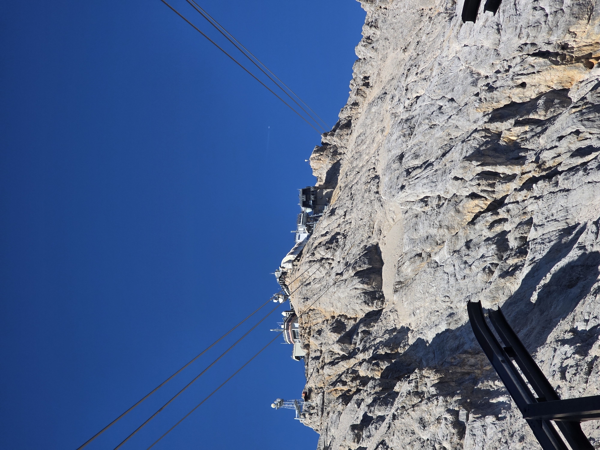 A low-angle, full shot captures the upper section of a rocky mountain peak under a clear, deep blue sky. Multiple thick cables stretch diagonally across the frame from the bottom left and right, disappearing towards the top. Perched on the summit are several buildings and structures, including what appears to be a ski lift station and a communication tower with antennae. The rugged, grey-brown rock face shows visible striations and shadows, indicating a sunny day. In the lower left corner, a dark, metallic structure, possibly part of the cable car infrastructure, enters the frame. No people are visible in the image. The scene is set at the Zugspitze, Germany's highest peak, located near Garmisch-Partenkirchen.