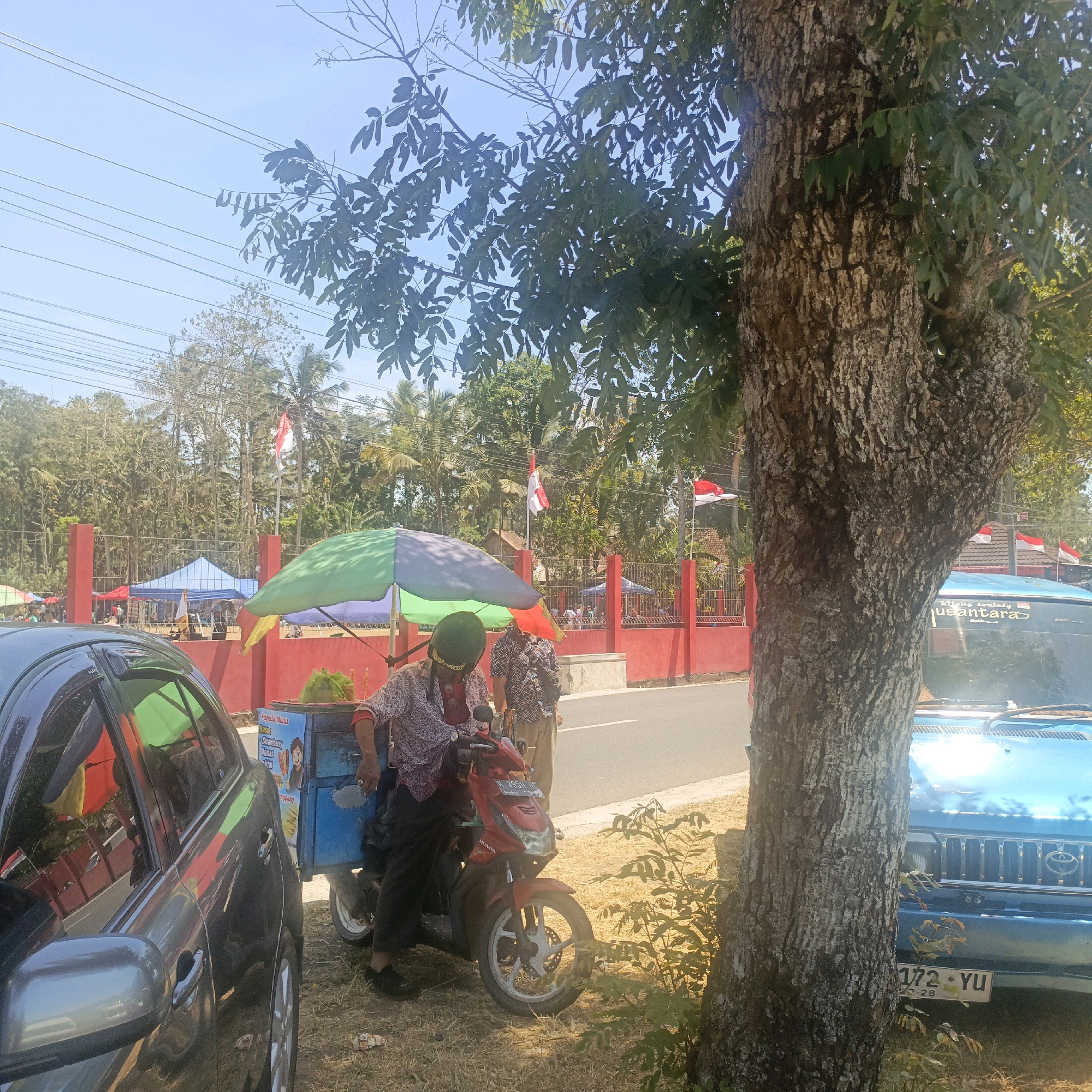A street scene in Krajan Lor, Indonesia, features a vendor on a red Honda scooter next to an ice cream cart. The vendor, wearing a patterned shirt and dark pants, is seated on the scooter, which is parked on dry grass next to a paved road. The ice cream cart is blue with a colorful umbrella providing shade. In the background, a red fence borders a street lined with trees, palm trees, and some buildings. Indonesian flags are visible on poles behind the fence. To the left of the vendor, a dark gray car is parked, and to the right, the front of a blue vehicle is visible, displaying a license plate that reads "172 YU". The sky is clear and blue, indicating a sunny day.