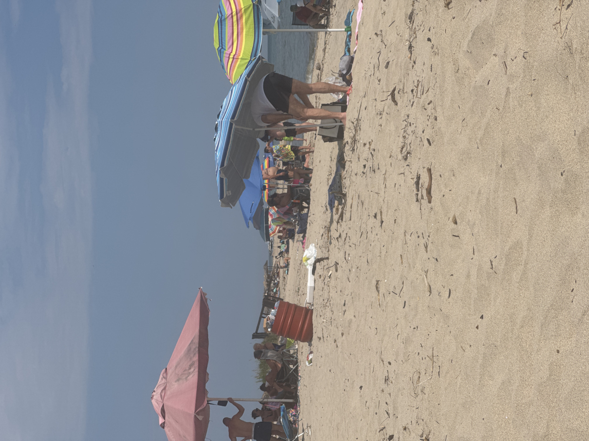 The image depicts an outdoor beach scene under a hazy blue sky, identified as the city of Unknown, Unknown. The upper approximately one-third of the frame shows a light blue sky with faint, diffuse cloud cover. The lower two-thirds are dominated by a tan sandy beach extending into the background, where the ocean is visible on the right horizon.
In the left midground, a large, faded pink beach umbrella provides shade. Beneath it, a shirtless man in dark shorts is visible, leaning against the umbrella pole. To his right and slightly behind him, other individuals are seated in beach chairs, partially obscured. Directly in front of this group, a weathered, ribbed red plastic barrel, likely a trash receptacle, stands upright, partially filled with discarded items. To the right of the barrel, a white styrofoam container and a crumpled white plastic bag are on the sand.
In the center-right midground, a person wearing a white t-shirt and dark shorts is bent forward, facing away from the viewer, appearing to retrieve or place an item into a dark tote bag situated on the sand. This individual is positioned under a blue and white striped beach umbrella. Immediately to their right, a multi-colored (yellow, green, pink, blue stripes) beach umbrella is open. Further back in the midground, numerous other beachgoers are visible, scattered across the sand, some under additional blue umbrellas, engaging in various activities such as sitting and standing.
The sandy foreground and midground are covered with numerous pieces of natural debris, including small sticks, driftwood fragments, and dried plant matter. The sand itself shows subtle ripples and variations in texture. Personal belongings such as towels and flip-flops are also visible near the umbrellas.