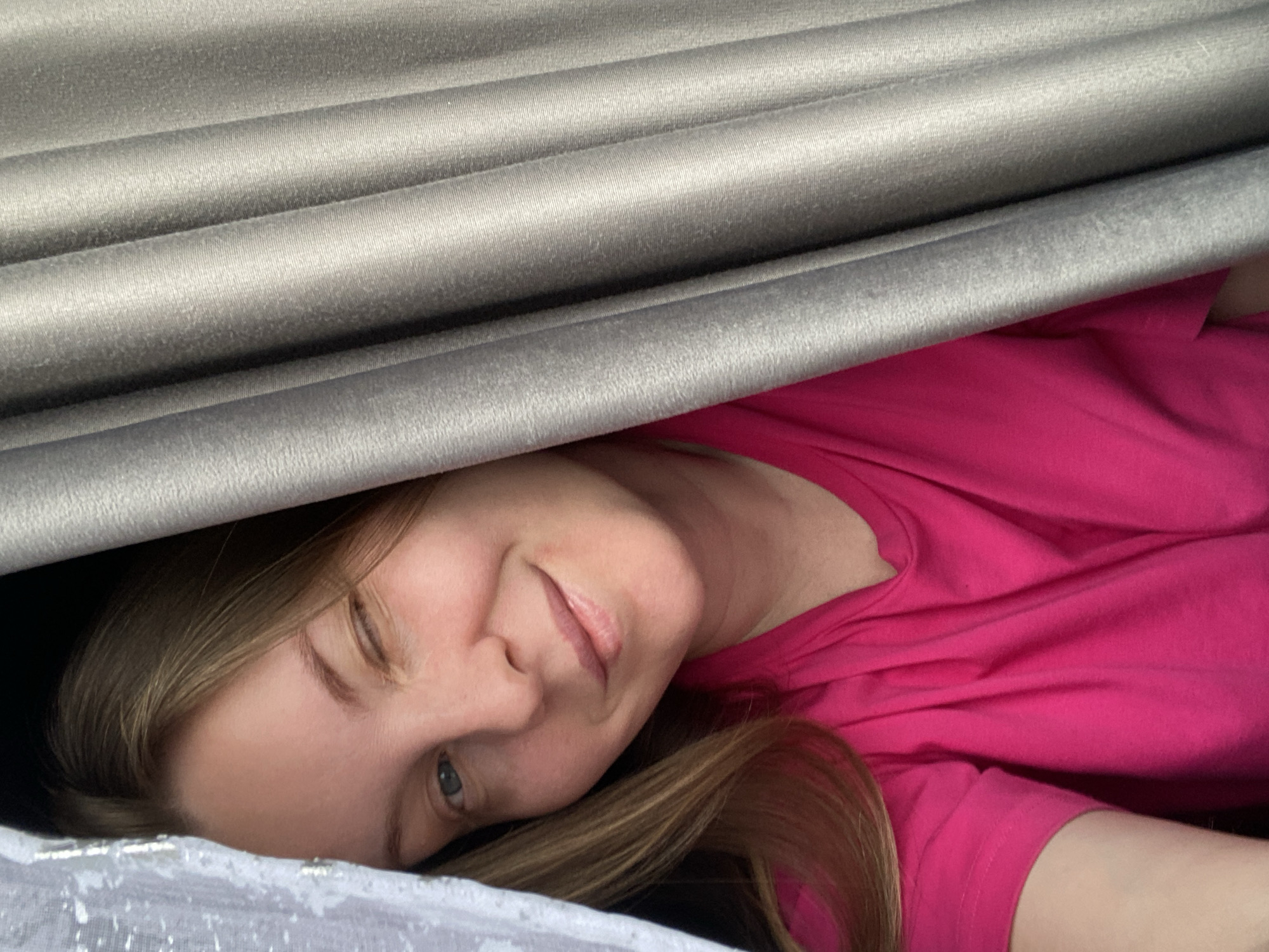 A fair-skinned woman with light brown hair lies on a white, textured surface while wearing a bright pink t-shirt. Her head is tilted, and she appears to be looking at the camera with a slight smile. Above her, a section of gray, corrugated material is visible. The location is unknown.