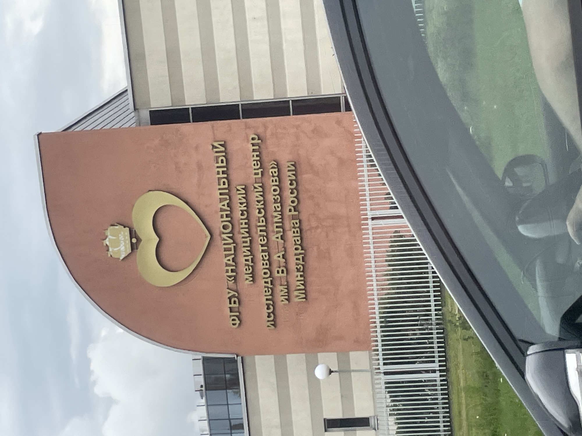 The image shows the entrance of the Almazov National Medical Research Center, located in Saint Petersburg, Russia. The building's facade features a large golden heart emblem topped with a crown. The text on the building reads "ФГБУ «НАЦИОНАЛЬНЫЙ МЕДИЦИНСКИЙ ИССЛЕДОВАТЕЛЬСКИЙ ЦЕНТР ИМ. В.А. АЛМАЗОВА» МИНЗДРАВА РОССИИ" which translates to "Federal State Budgetary Institution 'National Medical Research Center named after V.A. Almazov' of the Ministry of Health of Russia." The building has a distinctive curved section painted in a salmon pink color. To the right, part of the building is clad in beige and white horizontal stripes. A white picket fence runs along the front of the property, with a lamppost visible just behind it. The foreground is dominated by the reflection in the windshield of a car, showing a partial view of the car's interior and the exterior environment. The sky is overcast with clouds.