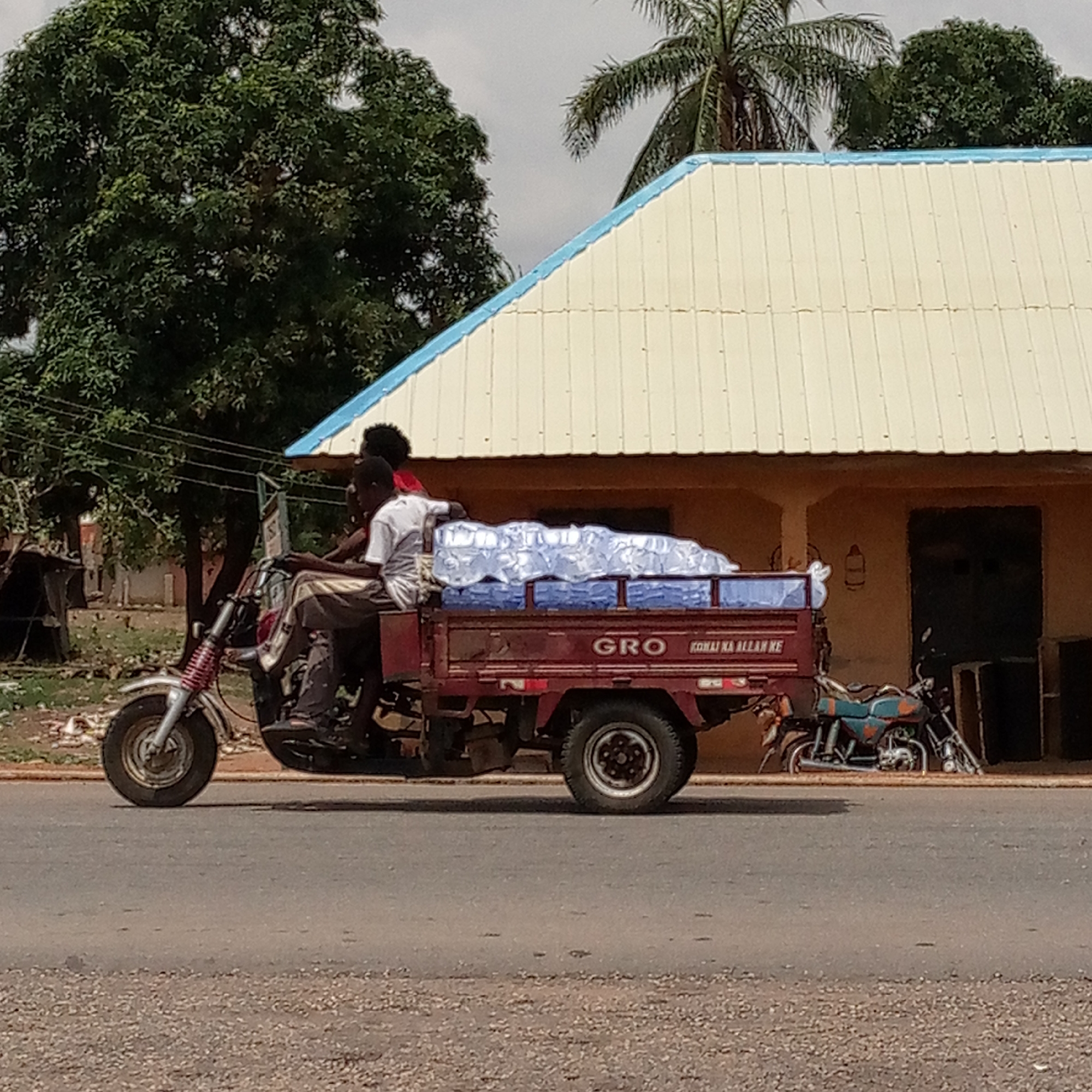 The image shows a three-wheeled motorcycle, commonly known as a cargo tricycle or mototaxi, being driven on an asphalt road during daylight hours. Two men are riding on the vehicle. The driver, wearing a light-colored shirt and dark trousers, is positioned at the front. A second man, wearing a red top, is seated behind him.
The tricycle's red cargo bed is heavily laden with numerous bundles of clear plastic bottles filled with water, indicating the vehicle is being used for transporting bottled water. On the side of the cargo bed, the text "GRO" and "KONAI KA ALLAH KE" is clearly visible.
In the background, there is a building with a light-colored, possibly cream or yellow, corrugated roof featuring a light blue trim along its edges. Behind and to the left of the building, a large, dense green tree stands, while a palm tree is visible further right, above the building. The sky appears overcast or hazy. To the far right, another motorcycle, painted blue and orange, is parked near the building. The ground in the foreground consists of asphalt with scattered small rocks or gravel. The scene depicts a typical daily activity in a semi-urban or rural setting, likely in a warm climate.