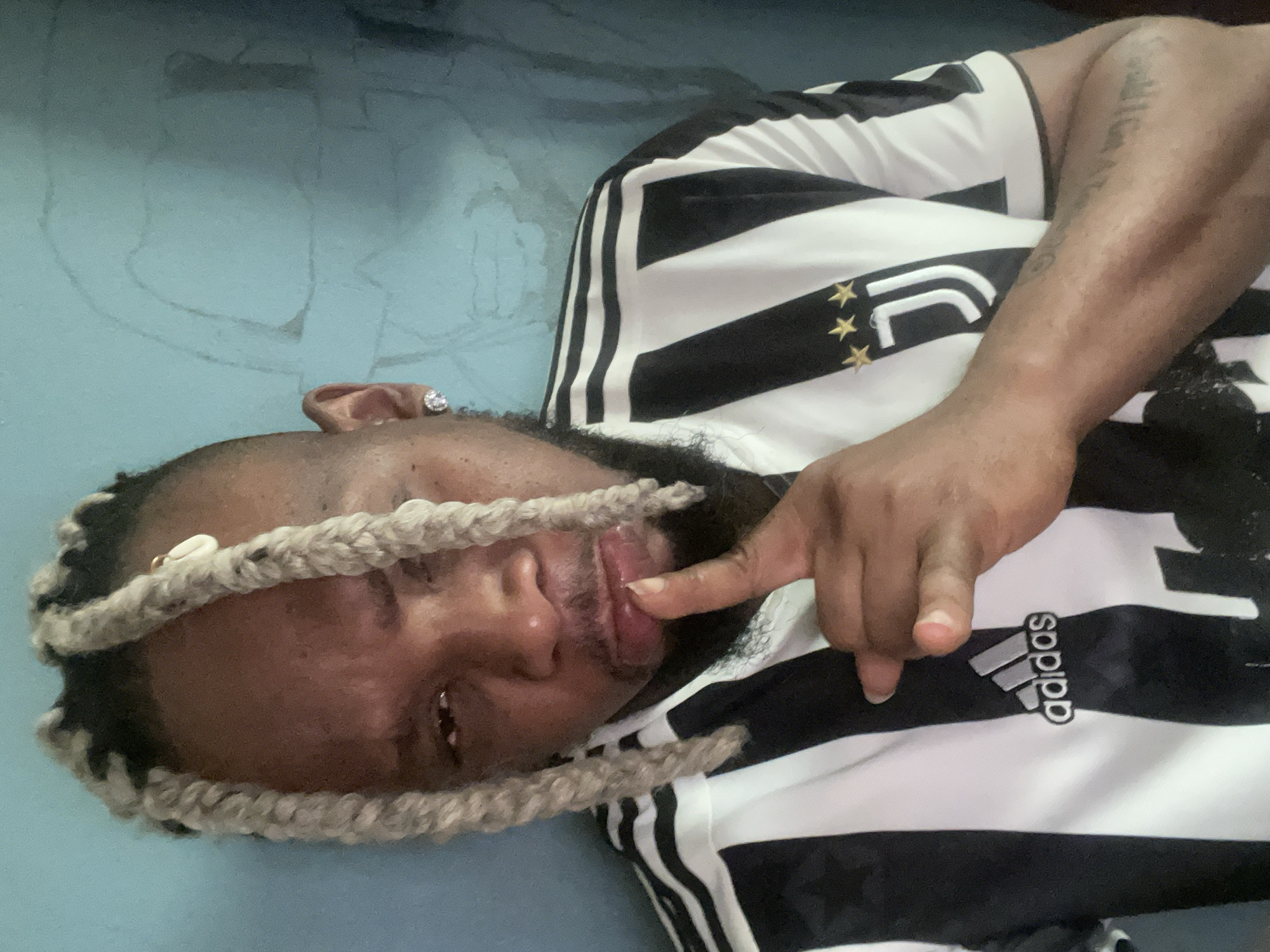 A close-up, chest-up portrait shows a Black individual with blonde, braided hair, wearing a black and white striped Adidas Juventus jersey. The individual is positioned facing the camera with their left hand raised to their lips, index finger pointed. They are wearing a diamond stud earring in their left ear. The background is a muted blue wall with faint, sketchy drawings. The image appears to be taken indoors, possibly in the city of Jos, Nigeria, given the individual's appearance and the context of the provided information.