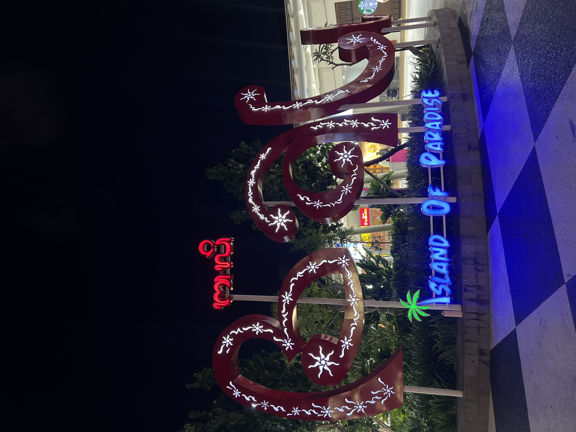 This night-time image captures a brightly lit, ornate sign display against a dark sky, likely marking an entrance or prominent location in Kuta, Indonesia.
The main subject is a large, elaborate red metal sign, styled with elegant swirls and heart-like shapes. It is adorned with numerous white, illuminated floral or starburst patterns that glow vividly. To its right, a blue neon-style sign spells out "ISLAND OF PARADISE" in capital letters, featuring a bright green palm tree icon positioned between "ISLAND" and "OF". Above and to the left of the large red sign, another red neon-style sign displays what appears to be Balinese script.
Behind these luminous signs, lush green foliage, including trees and bushes, is visible. In the upper background, parts of a well-lit building, possibly a shopping complex or mall, can be seen, with a recognizable partial Starbucks logo on one of its facades. The ground directly beneath and to the right of the signs features a distinct black and white (or dark and light grey) checkered pattern.
The image was taken at night, as indicated by the dark sky and the illumination of all the signs. There are no people visible, and the scene is static, showcasing decorative elements. The text "ISLAND OF PARADISE" combined with the palm tree and Balinese script serves as strong location cues for a tropical tourist destination, aligning with the provided context of Kuta, Indonesia. The overall atmosphere conveyed is one of welcoming vibrancy.