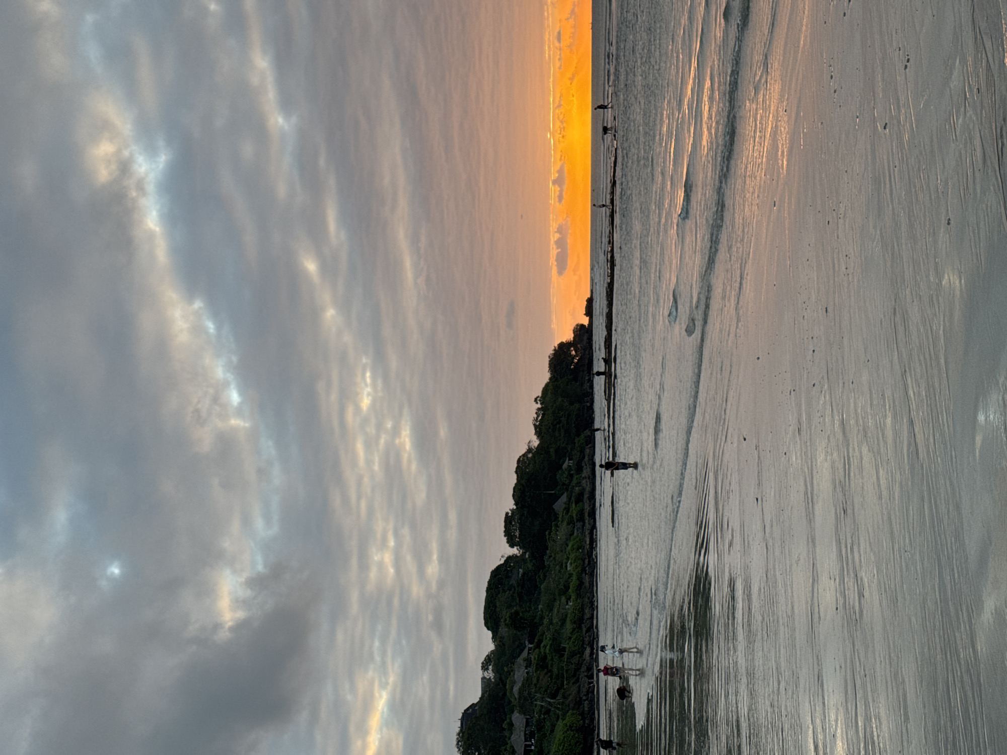 The image shows a beach scene at sunset in Kuta Selatan, Indonesia. The sky is filled with dramatic clouds, with a vibrant orange glow near the horizon where the sun is setting. Gentle waves lap onto the wet sand, reflecting the colors of the sky. Several people are visible on the beach, some walking near the water's edge, others standing in the shallow water. Lush green vegetation covers the hillside in the background, with some buildings nestled within it. The overall impression is one of a peaceful evening at the beach.