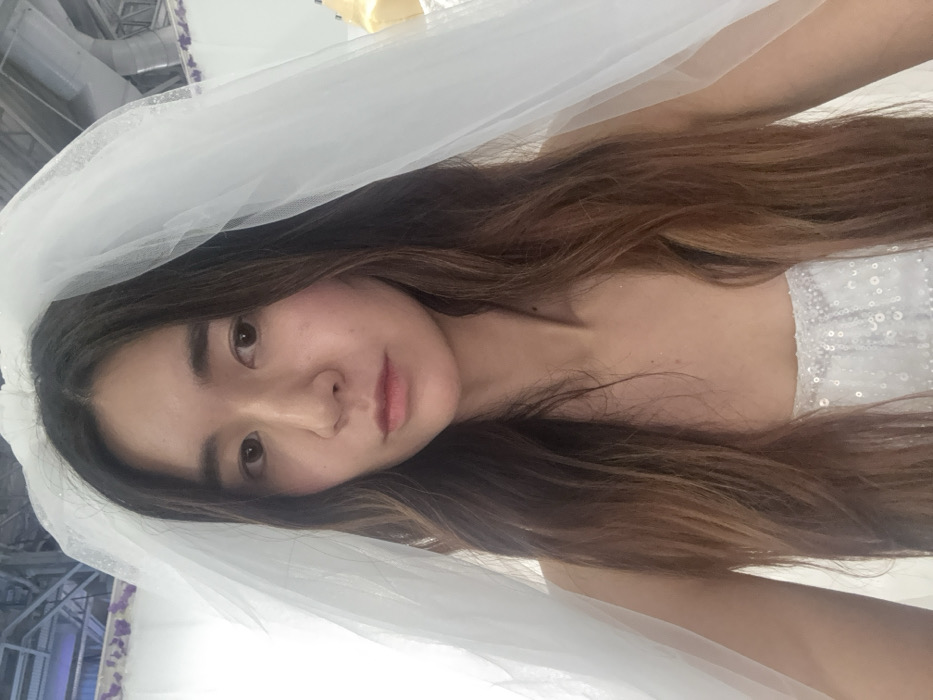 A woman with long brown hair is wearing a white wedding veil and dress. Her face is tilted slightly toward the camera. The dress appears to have some sequins or embellishments. The background is blurry, but suggests an indoor setting with some architectural elements visible.