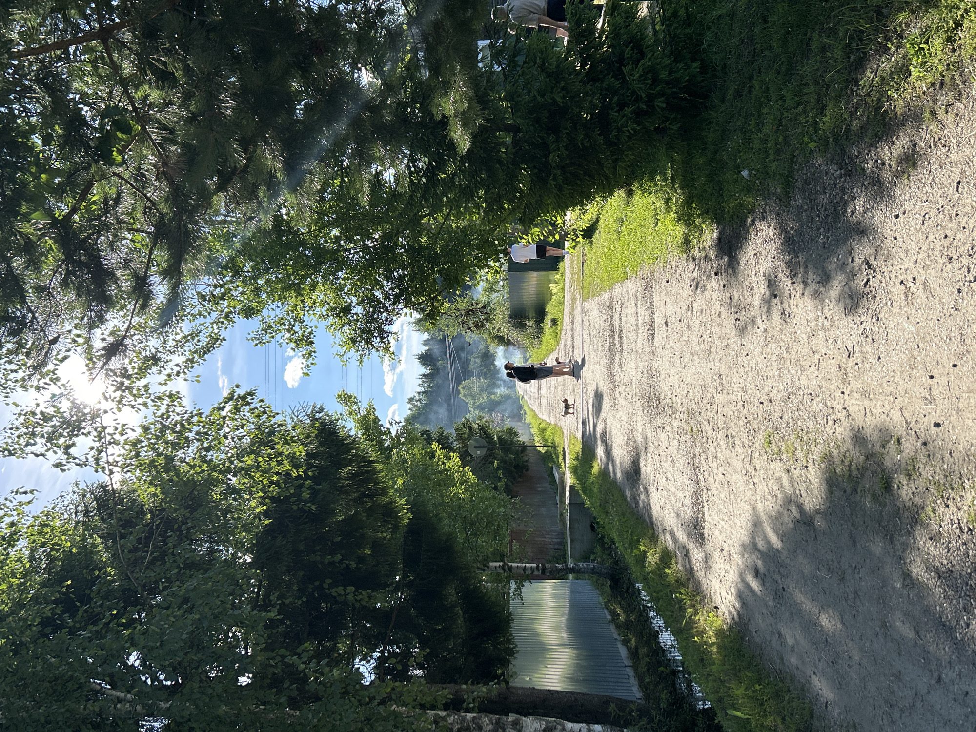 A dirt road stretches through a tree-lined area in Слава Снт, Russia. The sun shines brightly, casting shadows on the road. On the left side of the road, a corrugated metal fence runs along a ditch. On the right, a dense evergreen tree partially obscures the view of a house and other trees in the background.
Several people are present:
- Two individuals, one with dark clothing and another in white, walk together on the dirt road, accompanied by two small dogs.
- Further down the road, another person in white shorts and a white t-shirt walks away from the camera.
- To the far right, a person in a light-colored shirt stands near the evergreen tree.
The environment appears to be rural or suburban, with evidence of a residential area and natural vegetation. The sky is blue with some scattered clouds, and there is a faint haze or smoke visible in the distance.