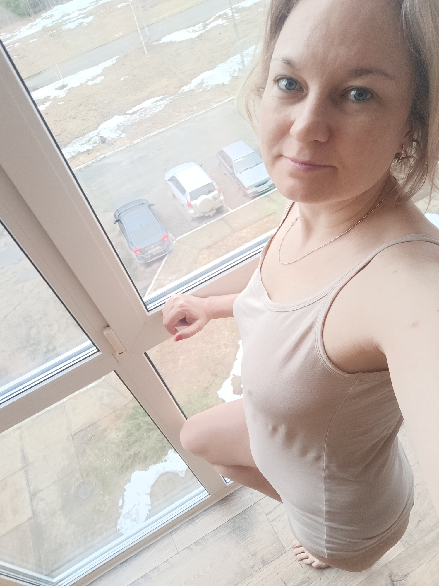 A woman with fair skin and light blonde hair is kneeling near a window. She is wearing a light beige tank top and a delicate silver necklace. Her expression is neutral, and she is looking directly at the camera. Outside the window, three cars are visible in a parking area, and patches of snow are present on the ground. The scene appears to be in a residential area in winter or early spring. Based on the location provided, the scene is in the city of Марковский, Russia.