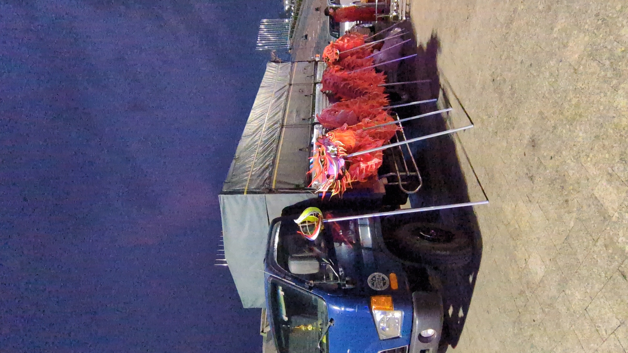 A blue truck is parked on a paved surface, adorned with multiple red Chinese dragon puppets mounted on poles. The truck bed is covered with a gray tarp. The setting is likely in a city, possibly Da Lat, Vietnam, given the presence of the Chinese dragons and the overall ambiance. A night sky provides the backdrop.