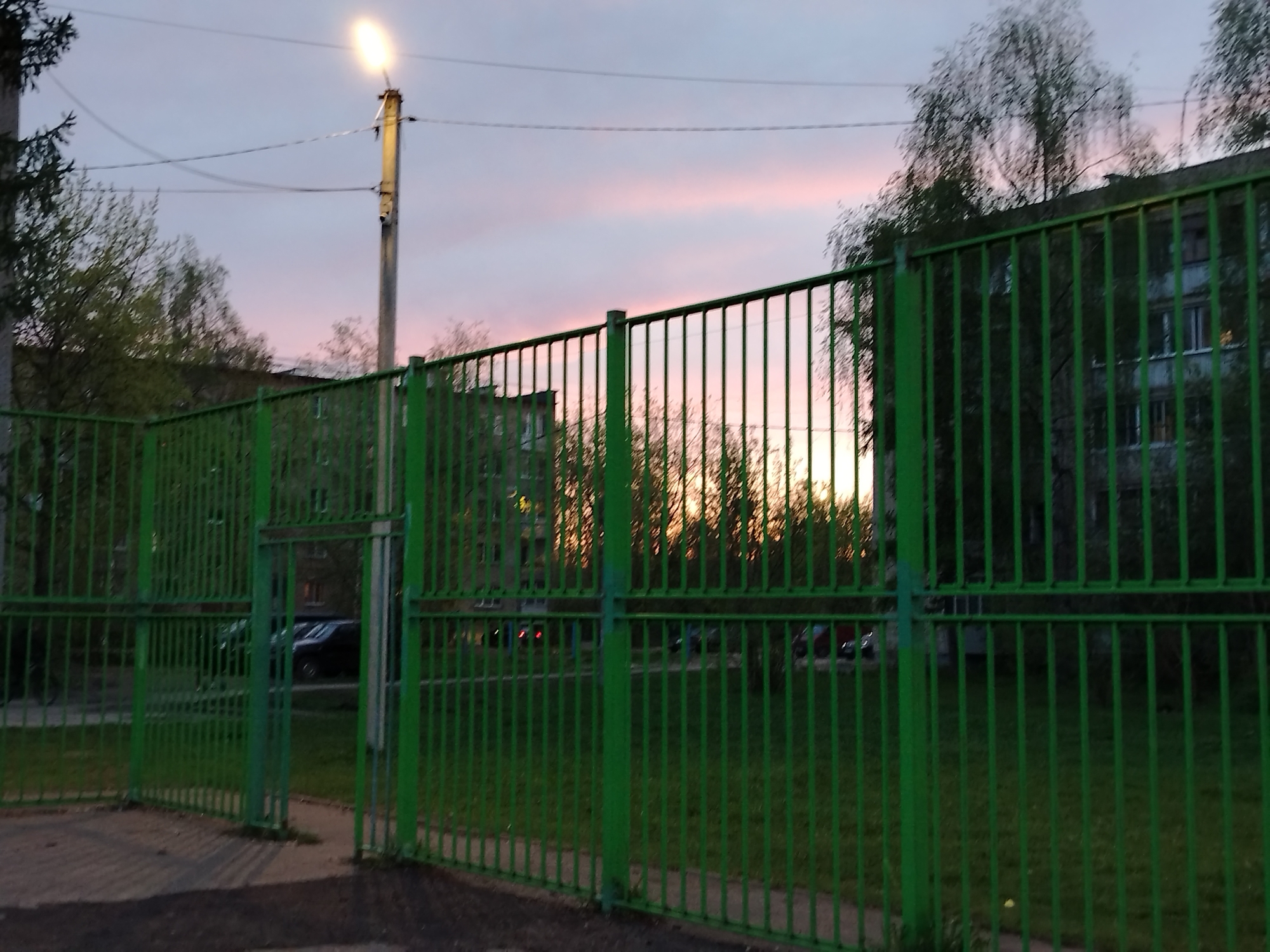 A green, metallic fence surrounds an area, partially obscuring the view of buildings and trees in the background. The sky above exhibits a transition from light pink to dusk colors. A streetlight is positioned on the left side of the frame, and cars can be seen through the fence. The location is Unknown, Unknown.