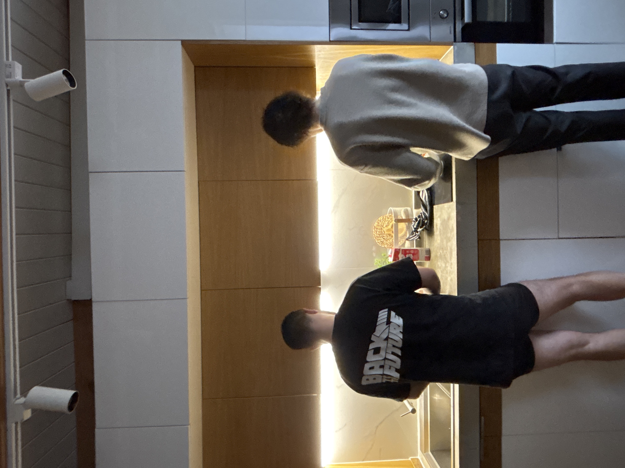 The image, oriented on its side, depicts two men in a modern kitchen in Chaikovsky, Russia. Both individuals are seen from behind, leaning over a dark kitchen counter that is brightly illuminated by a horizontal LED strip light installed under the upper cabinets.
The man positioned on the left wears a black t-shirt featuring the stylized text "BACK THE FUTURE" prominently displayed across his back, along with black shorts. He appears to be engaged in an activity at the counter, near a kitchen sink. The man on the right is dressed in a light-colored, long-sleeved top and dark trousers. He also leans over the counter, focusing on a task. Both men have dark, short hair.
The kitchen environment includes light-toned wooden lower cabinets and white upper cabinets. A built-in microwave or oven with a metallic finish is visible within the upper cabinetry. A wicker basket and some small containers are placed on the counter. On the far left of the frame, two white cylindrical surveillance cameras are mounted vertically on a light-colored, possibly paneled wall. The scene is lit by artificial indoor lighting, making the time of day indeterminate. No specific emotions can be discerned as the subjects' faces are not visible.
