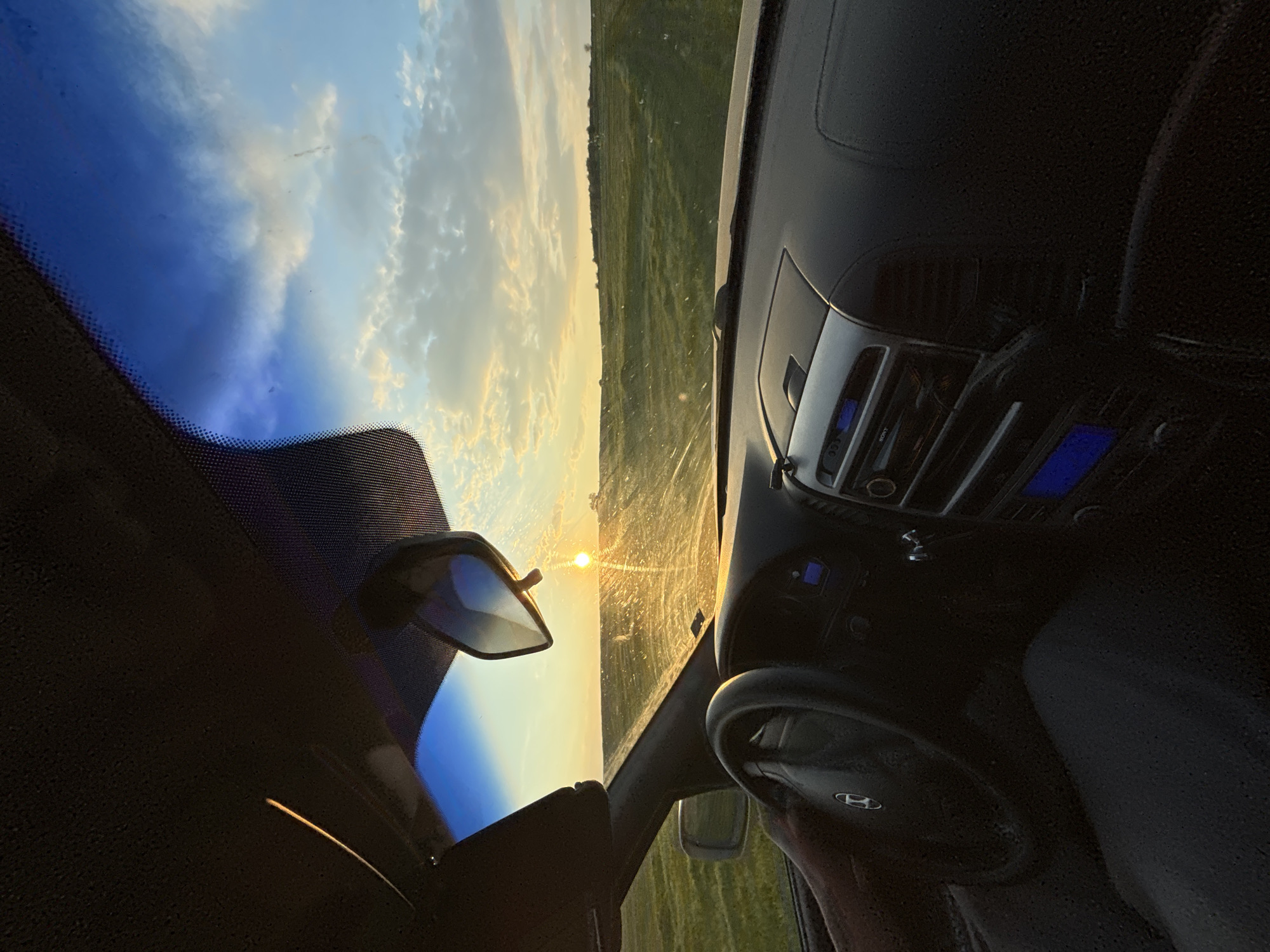 The scene is inside a car, likely a Hyundai, with a view through the windshield of a landscape with a setting or rising sun. The landscape appears to be fields, possibly in a rural area. The rearview mirror is visible. The image is rotated, with the horizon line appearing vertical. The location of Lehchevo, Bulgaria is not directly evident within the image. The time appears to be either sunrise or sunset based on the position of the sun.