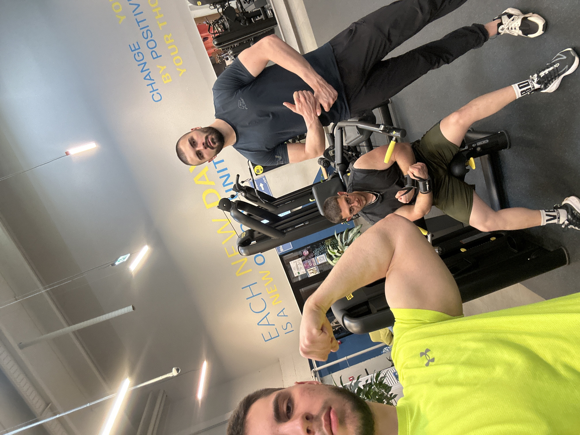 Three men are photographed inside a gym. One man in the foreground wears a neon green shirt and flexes his bicep for the camera. Another man is seated on a leg extension machine, wearing a black shirt and olive green shorts with black wrist wraps. A third man stands beside the seated man, wearing a dark blue shirt and black pants, making a hand gesture. Gym equipment is visible in the background. Wall text reads "EACH NEW DA IS A NEW O UNIT" and "CHANGE YO BY POSITIVE YOUR THO". The location appears to be a gym within the city of Sofia, Bulgaria.