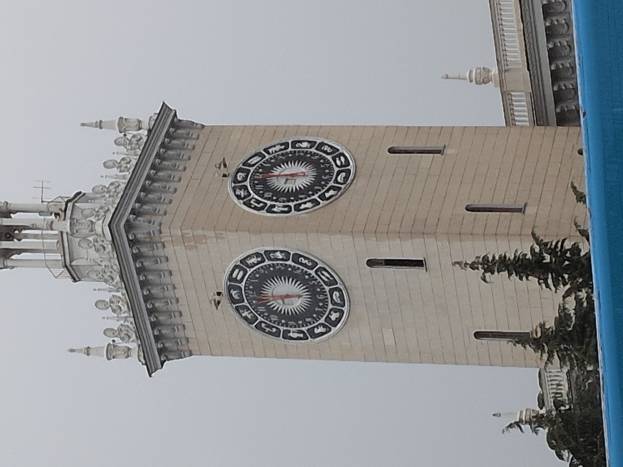 This is a medium-angle, eye-level, outdoor, daylight shot of the Sochi Sea Port clock tower. The sky is overcast and gray. The clock tower is a tall, rectangular structure made of light-colored stone blocks. It has decorative architectural elements at the top, including statues and finials. Two large clocks with astrological symbols are prominently displayed on opposite sides of the tower. The clock on the left has hands pointing to approximately 12:05, and the clock on the right also shows approximately 12:05. Below the clocks, there are narrow, vertical windows. In the foreground, the tops of evergreen trees are visible, and a blue structure, possibly a railing or a barrier, runs across the bottom of the frame. The architectural style suggests a Soviet-era design, common in port cities built during that period. The Sochi Sea Port is a well-known landmark in the city.