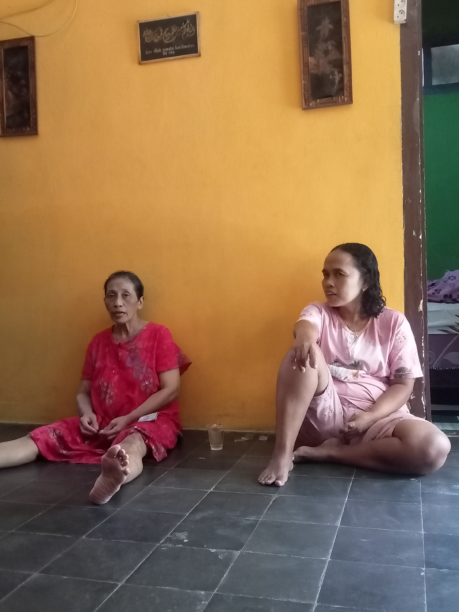 Two women are sitting on a dark tiled floor in front of a bright yellow wall. The woman on the left is older, with dark hair pulled back and is wearing a red floral-patterned dress. Her legs are extended in front of her. The woman on the right is younger, with dark, curly hair, and is wearing a pink pajama set with a floral pattern. She is sitting with her legs crossed. A small glass of liquid is on the floor near the woman in red. On the yellow wall, there are two framed pictures. The picture on the left is a dark, rectangular frame with an image inside. The picture on the right is a dark, rectangular frame containing Arabic calligraphy, with some text below it that appears to be in Latin script. The lighting suggests it is daytime.
