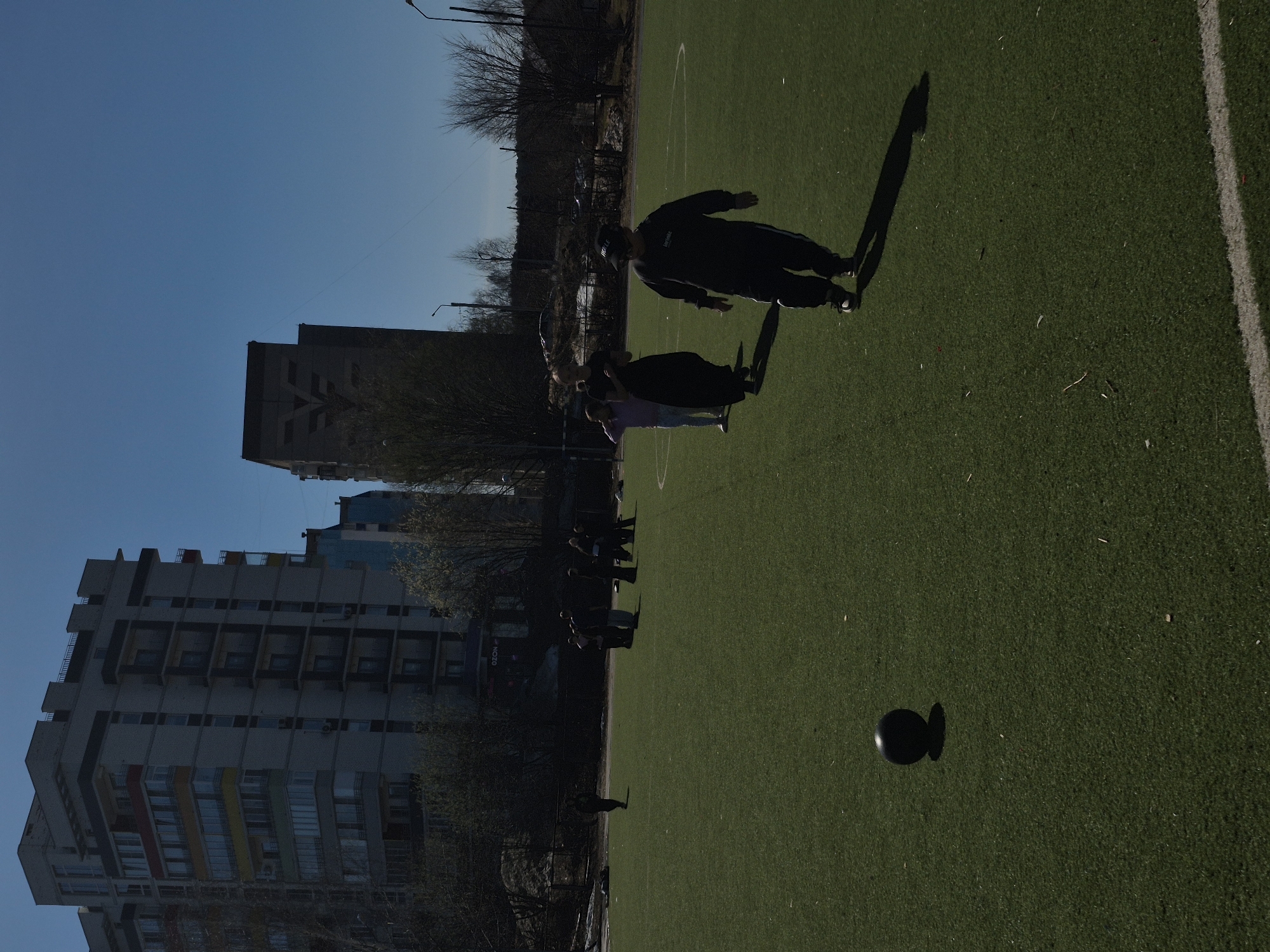 The image shows an outdoor scene in the city of Chaikovsky, Russia, during daylight. The location appears to be an artificial turf sports field, likely for soccer or football. On the left, several apartment buildings, typical of Soviet-era architecture, stand against a blue sky. The field is mostly green with white boundary lines. A group of people is present in the middle of the field. One individual wears dark clothing and appears to be engaged in a kicking motion. Shadows indicate that the sun is positioned relatively low. A dark, round object, possibly a ball, rests on the field near the bottom. The overall impression is of a recreational activity taking place in an urban setting.