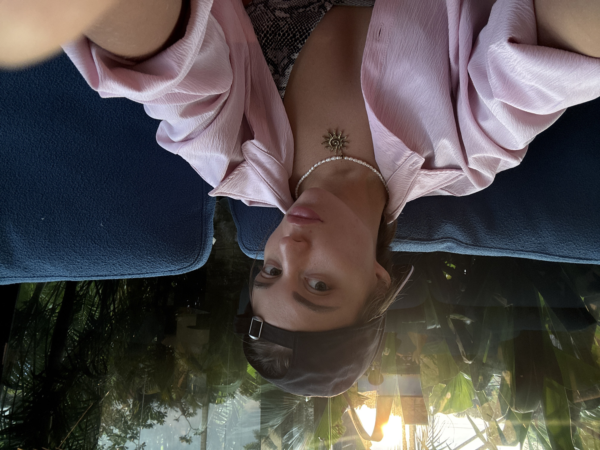 The provided image is an inverted, close-up portrait of a person, likely female, situated in what appears to be an indoor or sheltered setting with an outdoor view. The subject looks directly into the camera with a neutral expression.
She wears a light pink, textured long-sleeved shirt, unbuttoned to reveal a black and white patterned (possibly snakeskin) top. Adorning her neck is a pearl choker necklace featuring a golden, intricately designed sun-shaped pendant. A dark baseball cap is worn backwards, with its brim positioned away from the camera. Blue textured fabric, likely upholstery, frames the top and bottom of the composition.
The background, visible as a reflection or through glass, showcases lush green tropical foliage, including large palm-like leaves. A prominent golden light source, characteristic of either sunrise or sunset, is reflected in the lower right portion of the background, casting a warm glow. The tropical elements align with the given location context of North Kuta, Indonesia. There is no discernible text in the image.