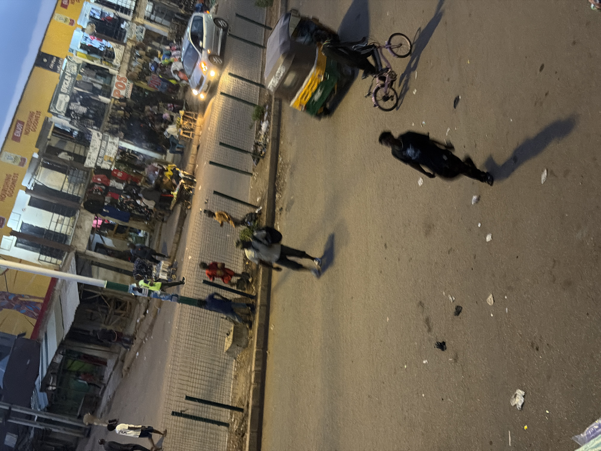 The image depicts a street scene in Bukuru, Nigeria, likely during the evening. Several individuals are present on the paved road and the sidewalk adjacent to a row of shops. On the sidewalk, a person in a yellow vest walks past a stall filled with clothing. Further down the sidewalk, other individuals are visible, some appearing to be shopkeepers or customers.
On the road, a person in dark clothing is walking towards the left side of the frame, casting a long shadow. To the right of this individual, a pink bicycle with a person on it is moving. In the background, a three-wheeled vehicle, commonly known as an auto-rickshaw or Keke Napep, is parked or moving slowly. A silver car is also visible on the road, its headlights illuminated.
The shops on the side of the street appear to be selling various goods, including clothing and other items. The environment suggests a busy marketplace or commercial area. The ground is littered with some debris. The lighting suggests it is either dusk or early evening, with artificial lights from the shops and vehicles contributing to the illumination.