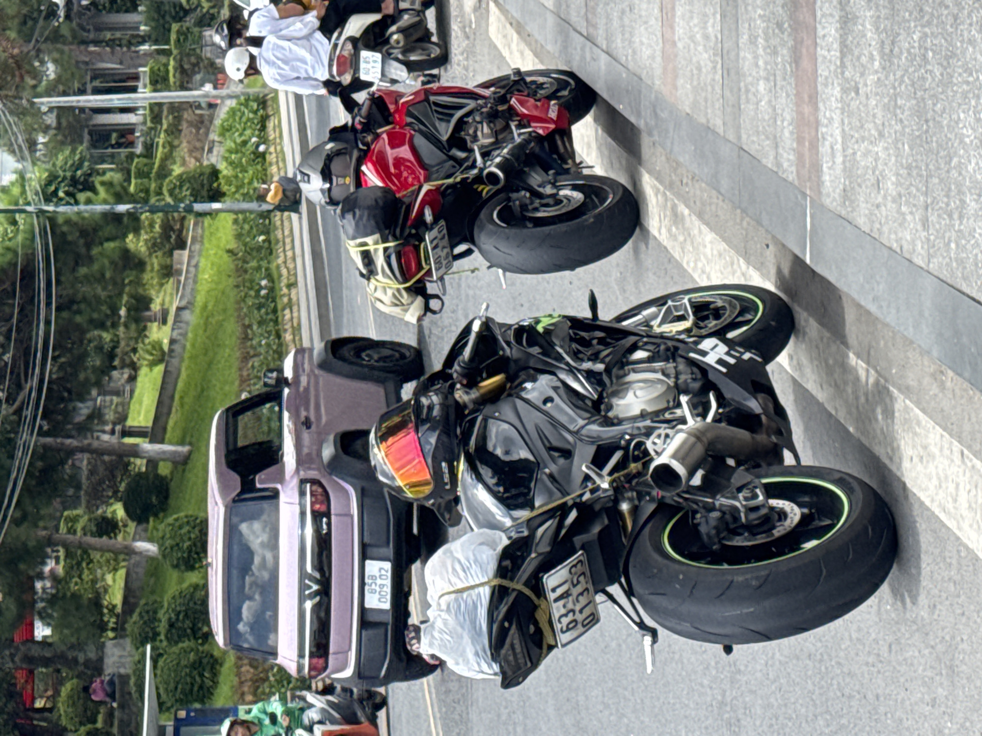 In Da Lat, Vietnam, on a paved road beside a sidewalk with green foliage and trees, two motorcycles are parked. The motorcycle in the foreground is black with a white plastic bag secured to its rear seat by ropes. A red motorcycle is parked behind it, also with luggage strapped to its rear. A purple SUV is visible in the background, with its rear facing the viewer. There are a few people in the image, including a person on a motorcycle to the left and two people on a motorcycle in the far right background. The scene appears to be a roadside in a city.